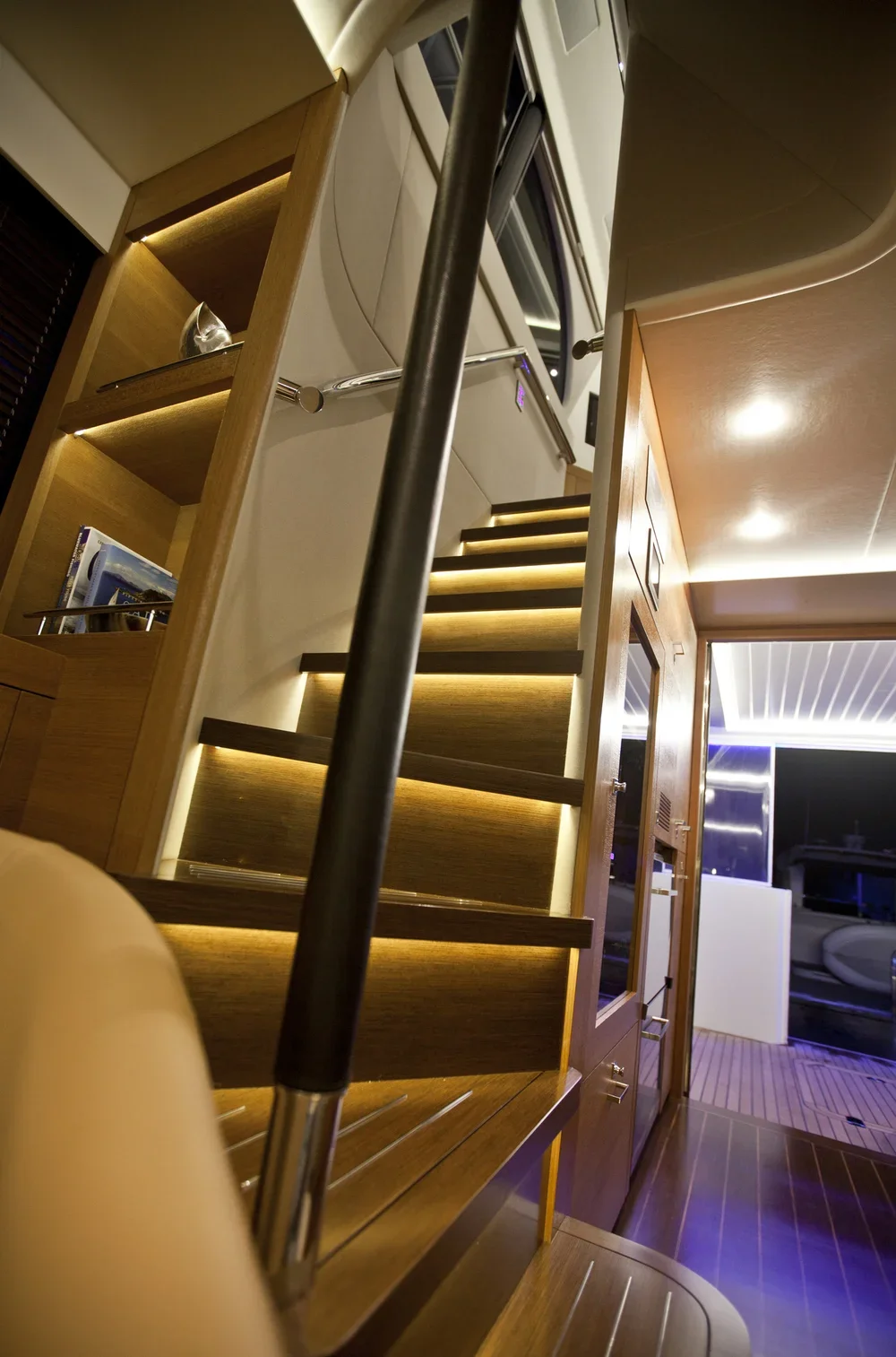 Interior view of a modern yacht or boat featuring a staircase with warm lighting, wooden shelves with books, and a glass door leading outside.