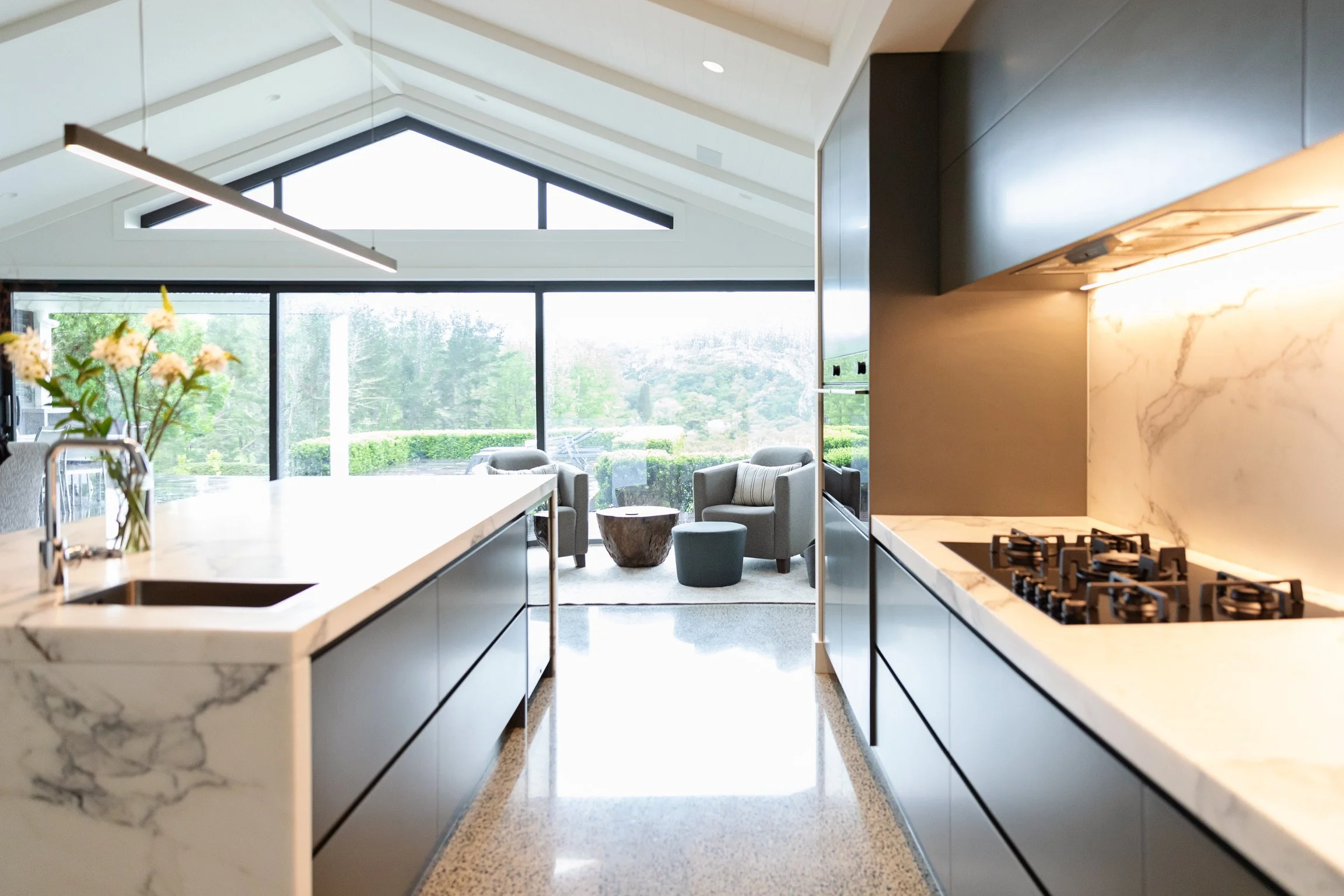 Modern open-concept kitchen with white marble countertops, black cabinets, and gas stove, leading into a living space with large glass windows, outdoor greenery, and seating area