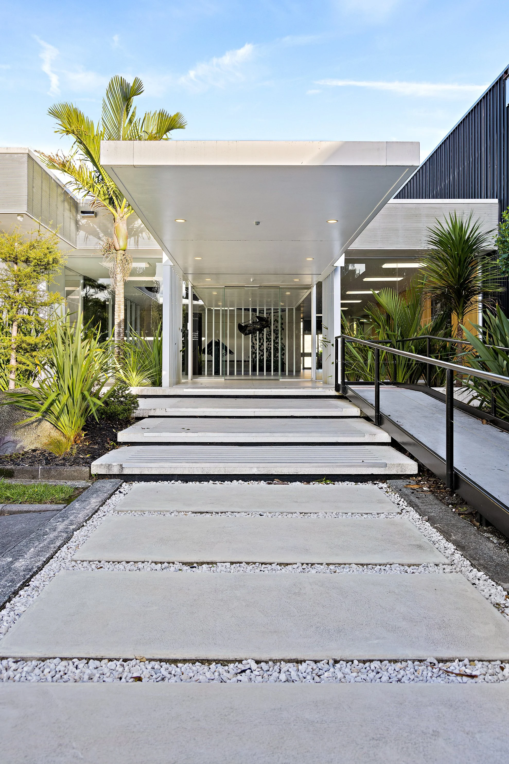 Modern building entrance with concrete steps, black railings, glass doors, and greenery including palm trees and shrubs.