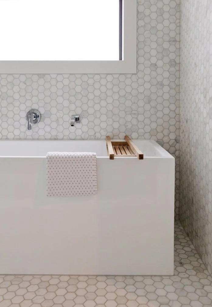 Bathroom with a white bathtub, hexagon tile walls, and floor. A towel hangs over the edge of the tub, and a small wooden bath tray rests on the tub edge. A window is above the tub.