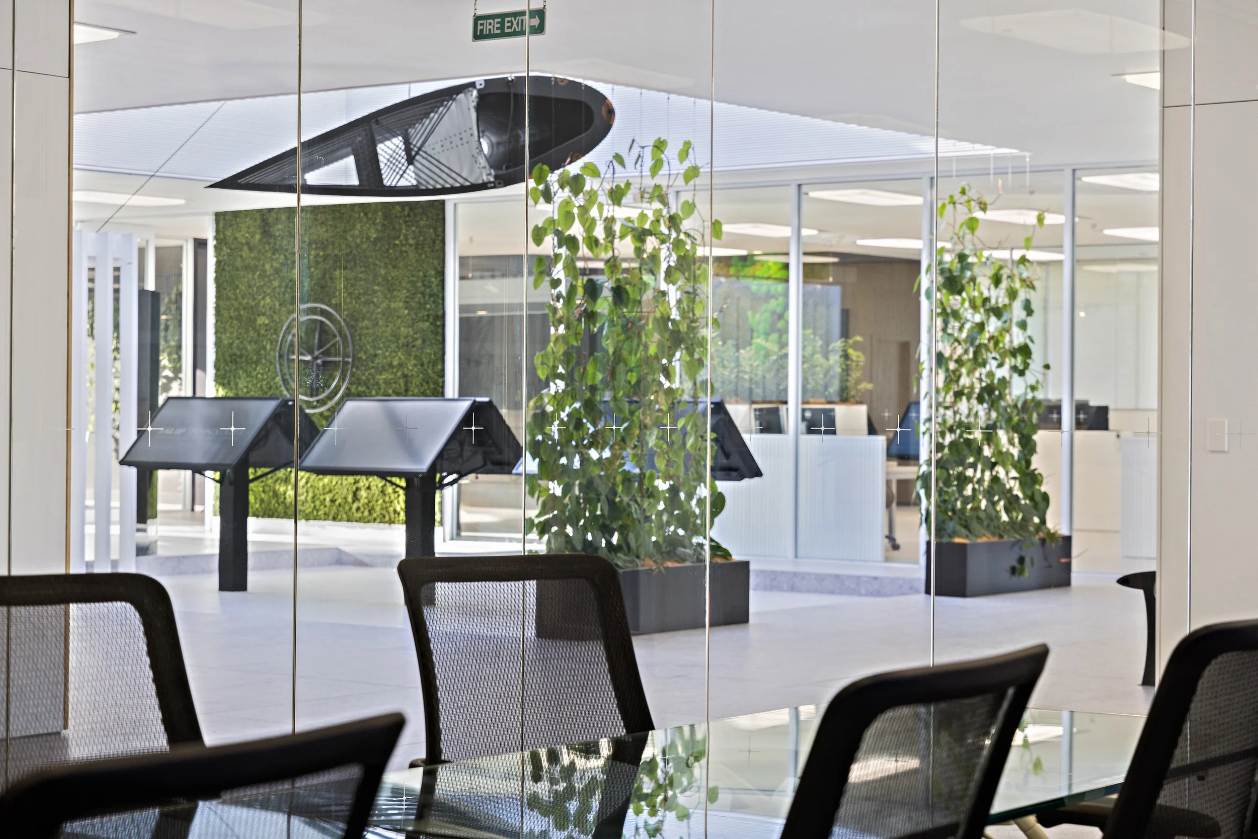 Interior of a modern office or lobby with glass walls, potted plants, chairs, and a clock, seen through glass with a reflection of chairs and tables.