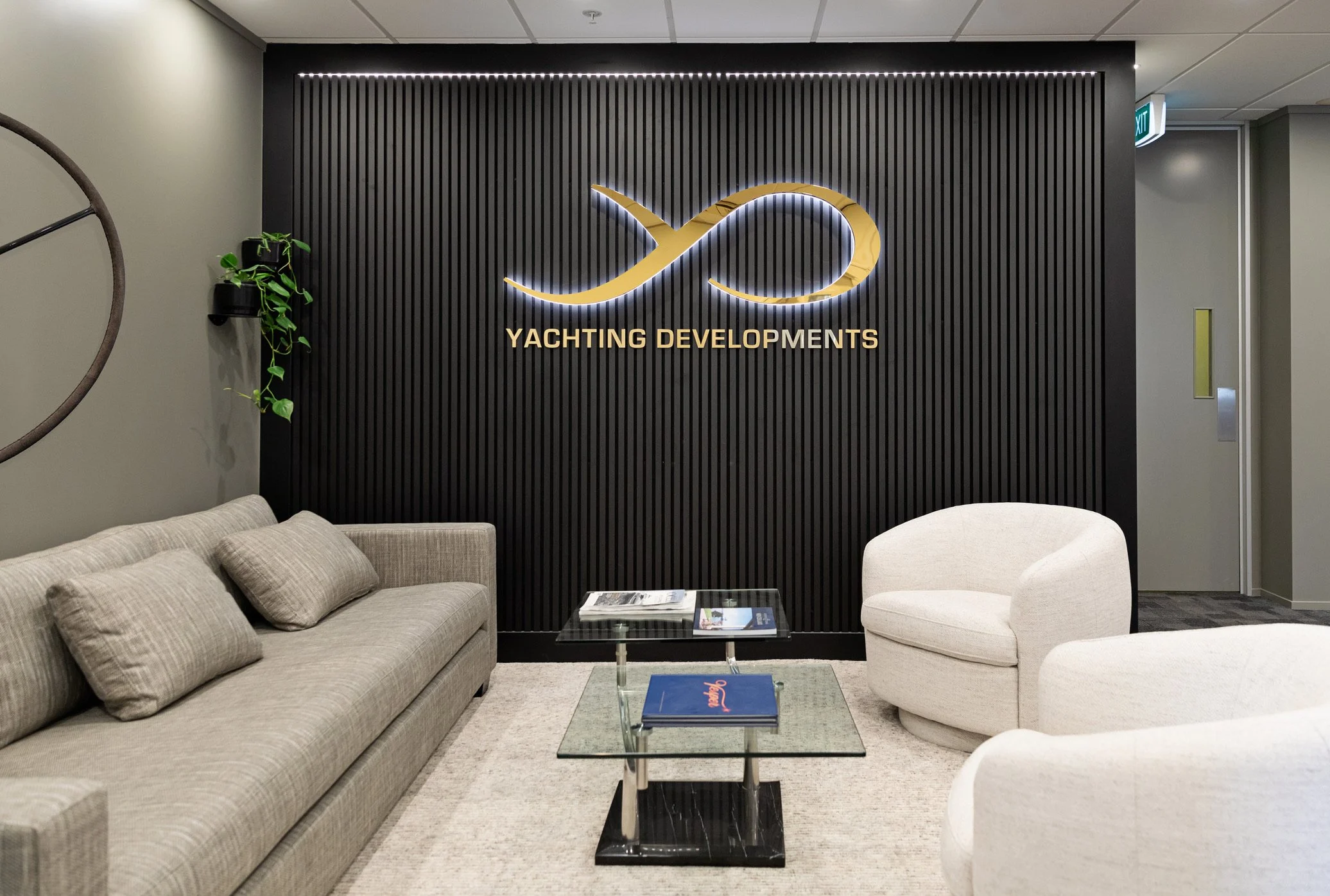 Office lounge area with beige sofa and white armchair, black slatted wall with illuminated infinity symbol and text 'Yachting Developments', glass coffee table with magazines