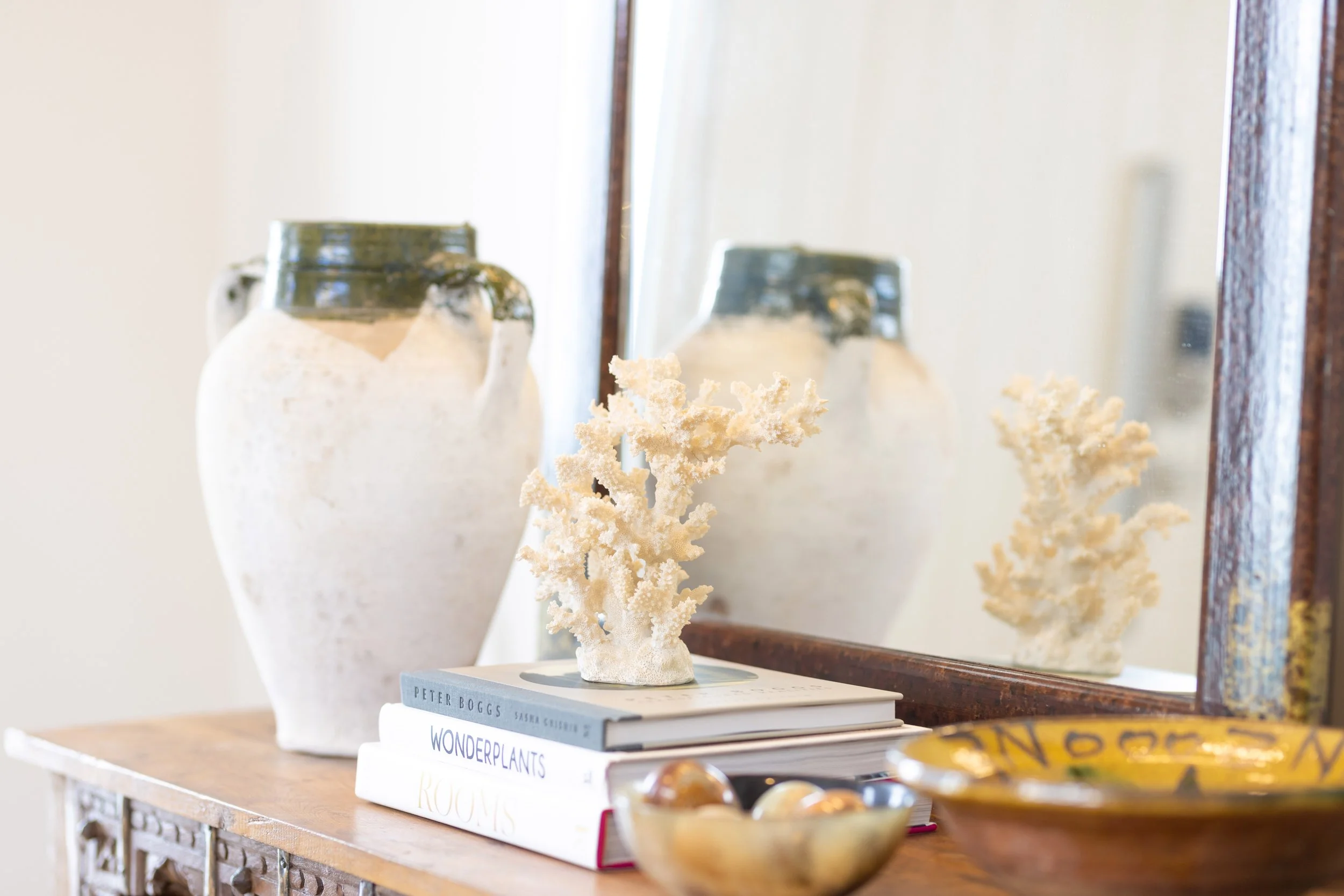 A decorative display featuring a white ceramic vase, a plant sculpture, and a stack of books on a wooden surface in front of a mirror.