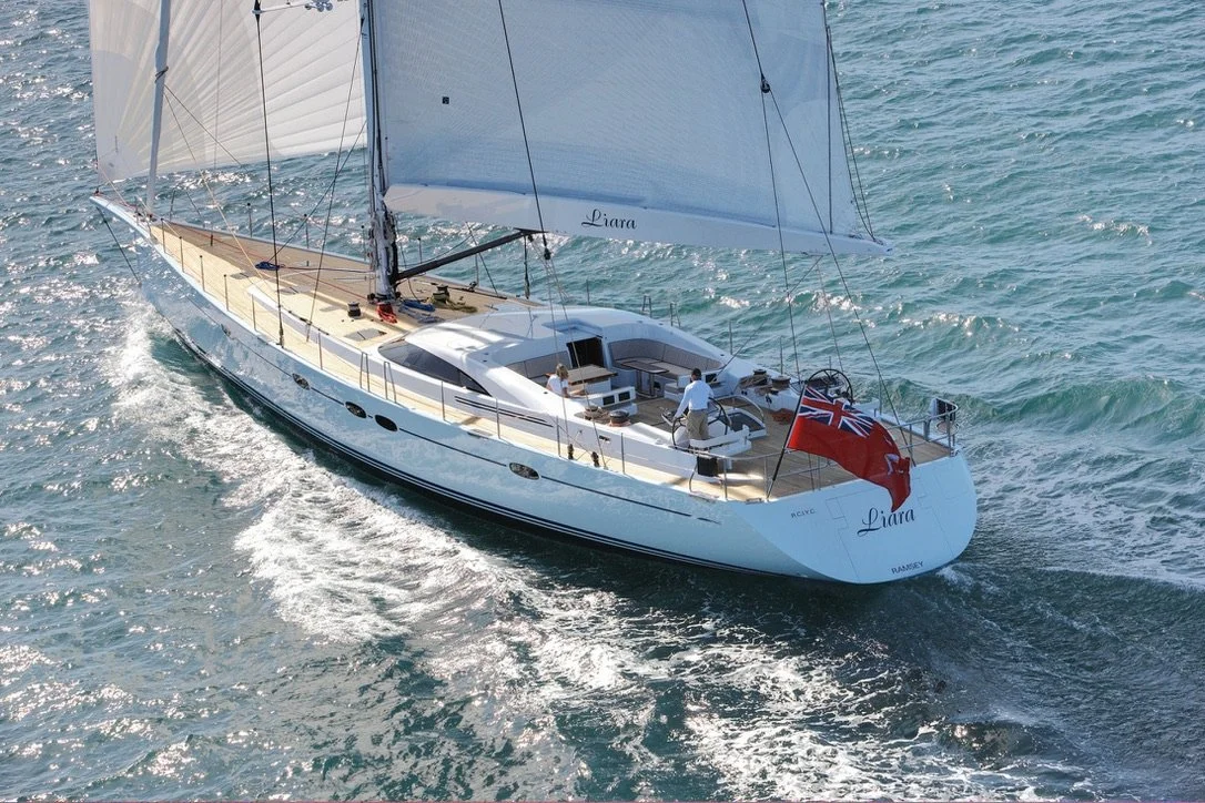 A white sailing yacht with two large sails, named 'Liara,' sailing on the water. The yacht has a couple of people aboard, and a British flag at the stern.