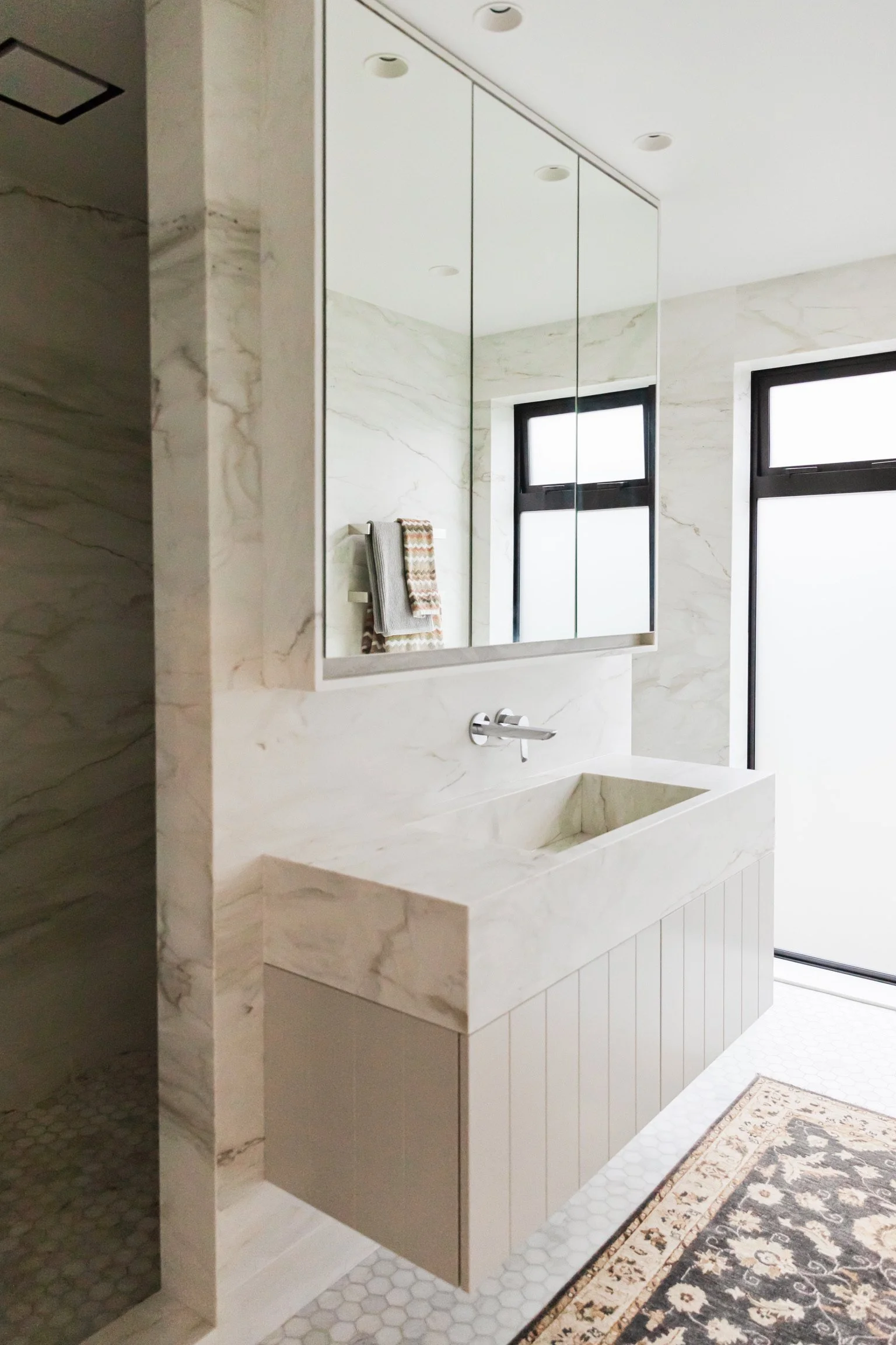 Bathroom with marble walls, a large mirror, a black framed window, a towel rack with two towels, a white washbasin, and a patterned rug on hexagonal tile flooring.