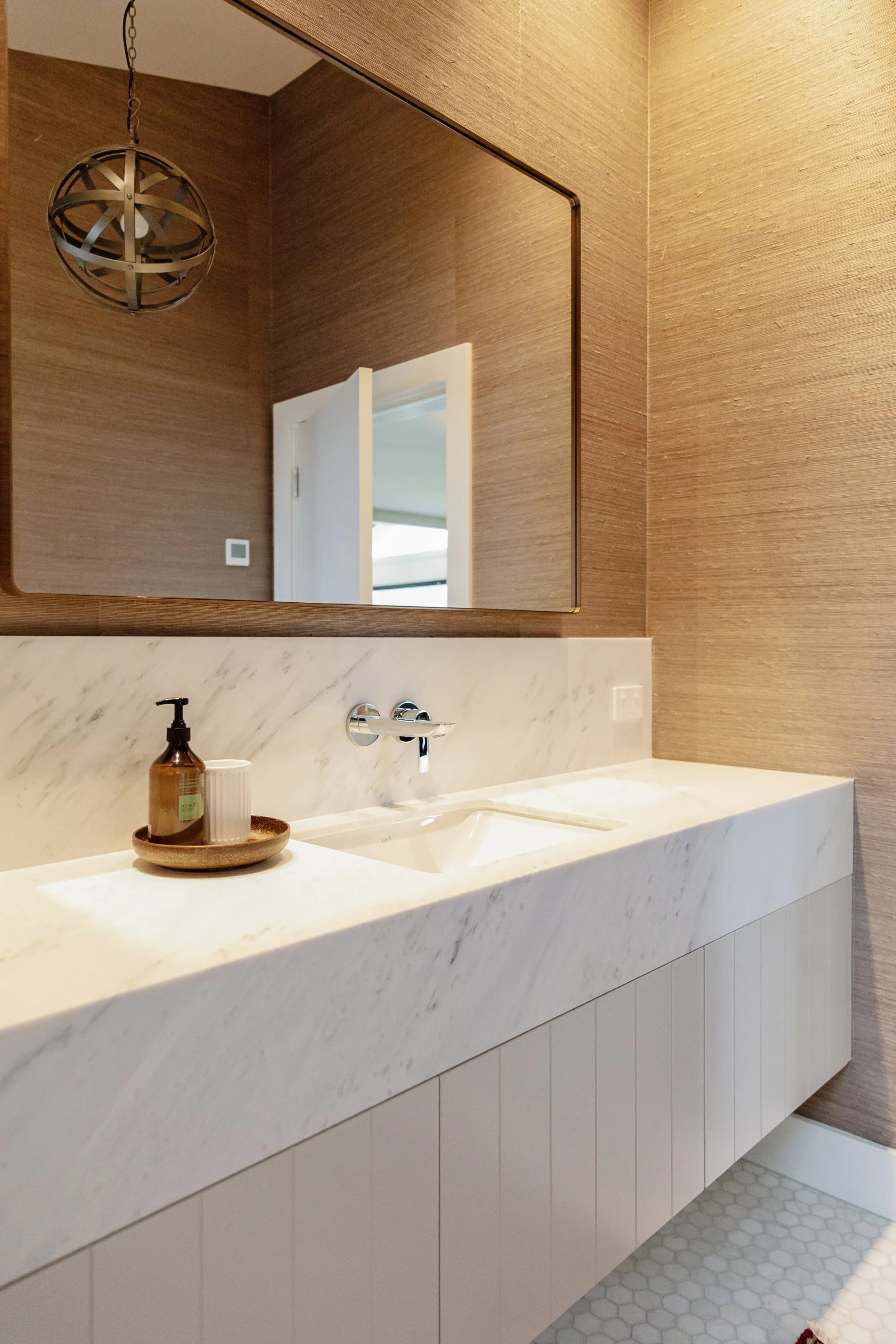 Bathroom vanity with a marble countertop, a rectangular sink, and a wall-mounted faucet. On the counter is a brown tray holding a brown soap dispenser and a white toothbrush holder. A large wall mirror reflects part of the room, including a door and 
