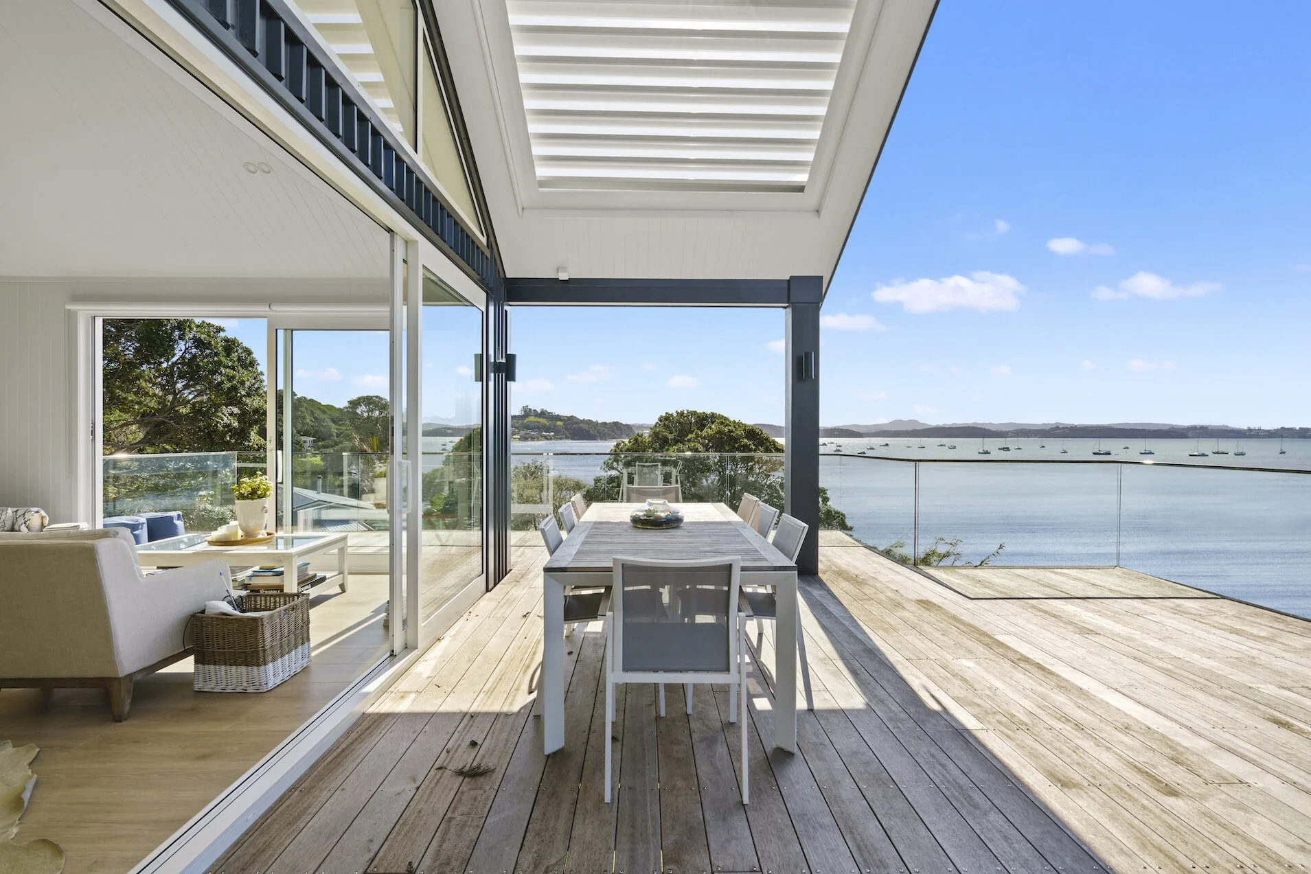 Modern outdoor terrace with wooden flooring, white dining table and chairs, and a view of a water body with boats and distant hills under a blue sky.