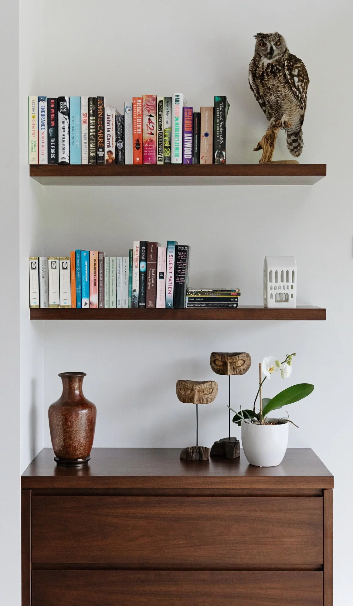 Wooden shelves with books, owl sculpture, white vase, decorative wooden masks, and orchid plant.