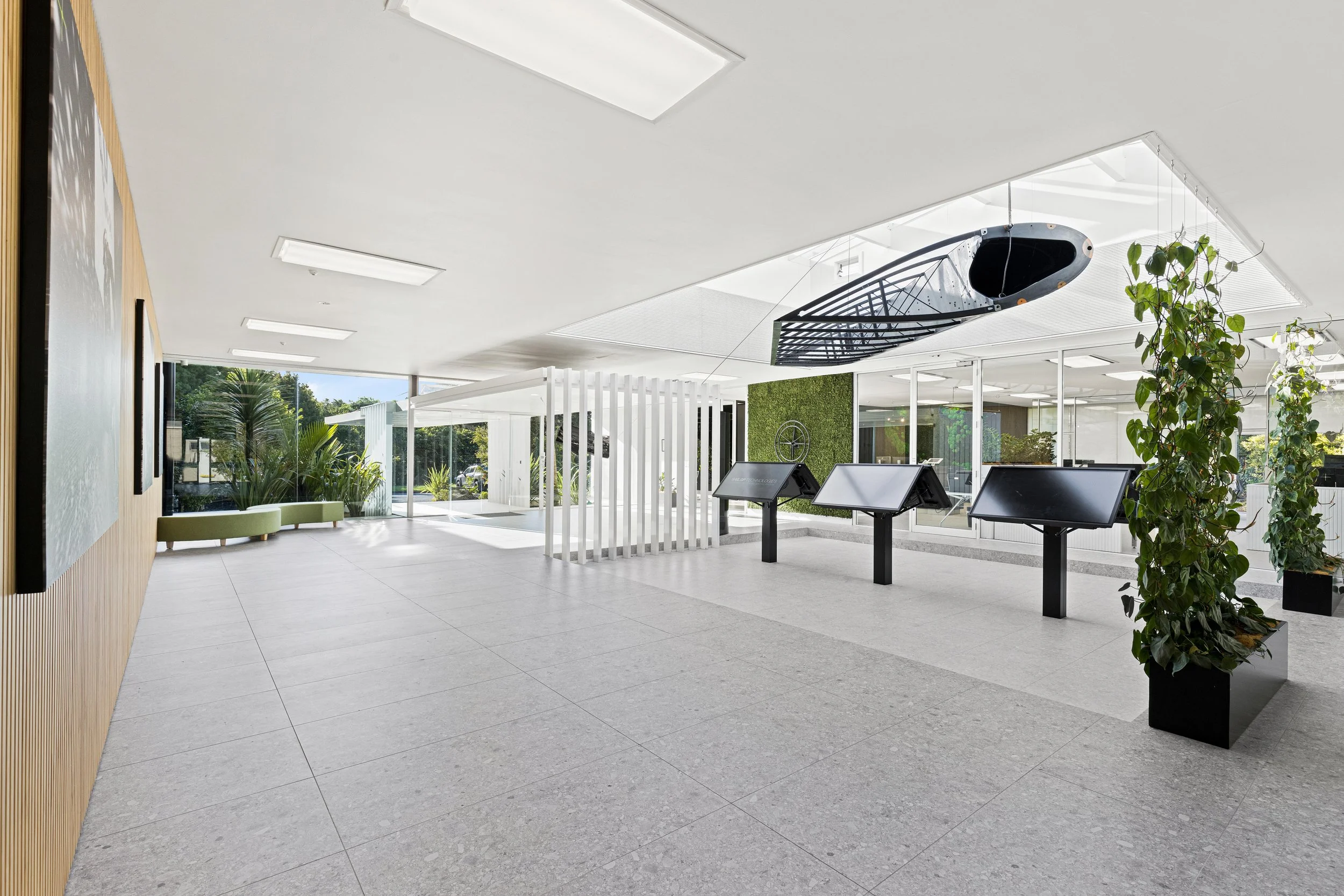 Modern indoor space with large windows, potted plants, digital kiosks, and a ceiling-mounted boat-shaped sculpture.