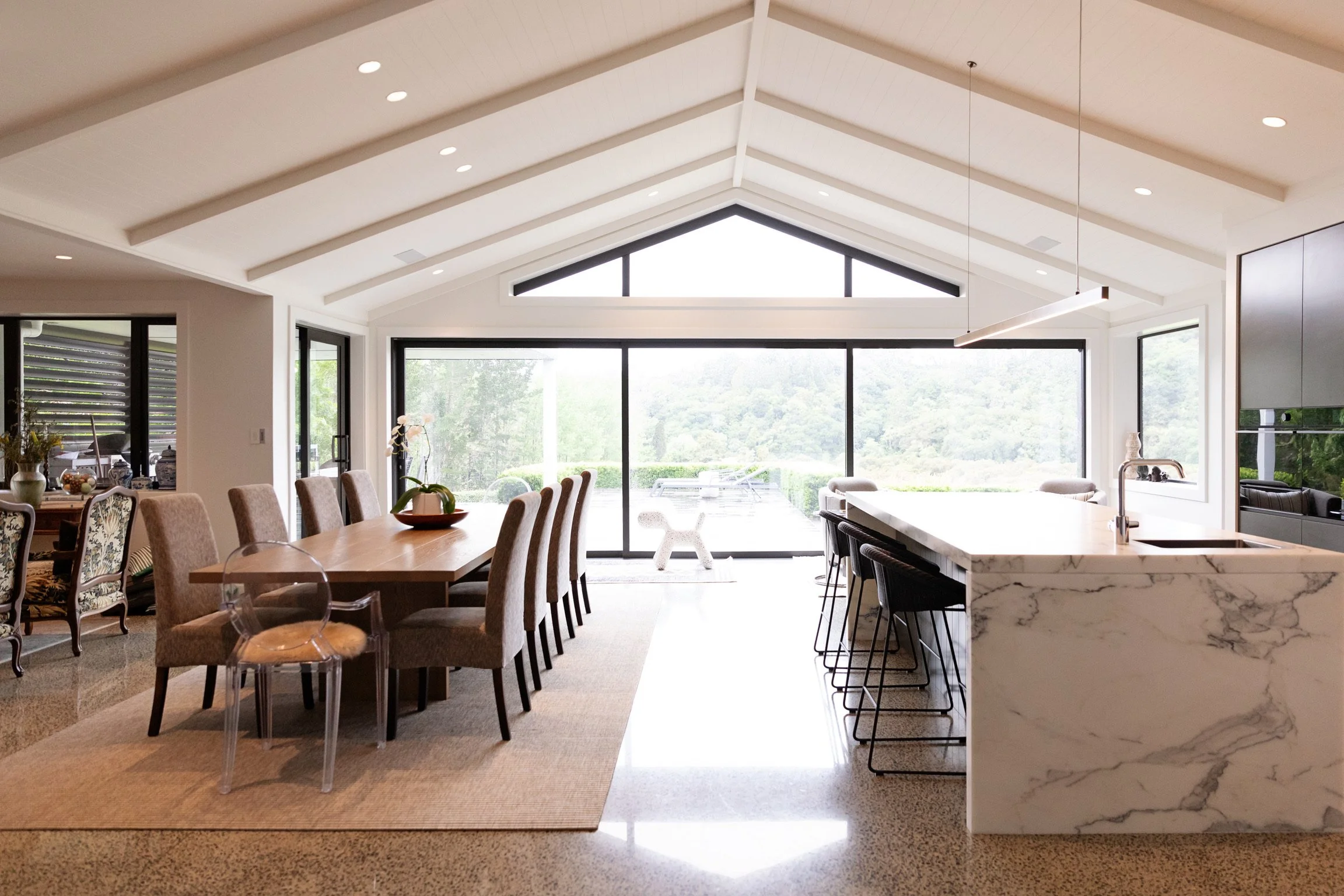A bright, modern living space with large windows showing a scenic outdoor view. The room features a dining area with a wooden table and upholstered chairs, and a kitchen island with a marble countertop and black bar stools. White walls and a high, va