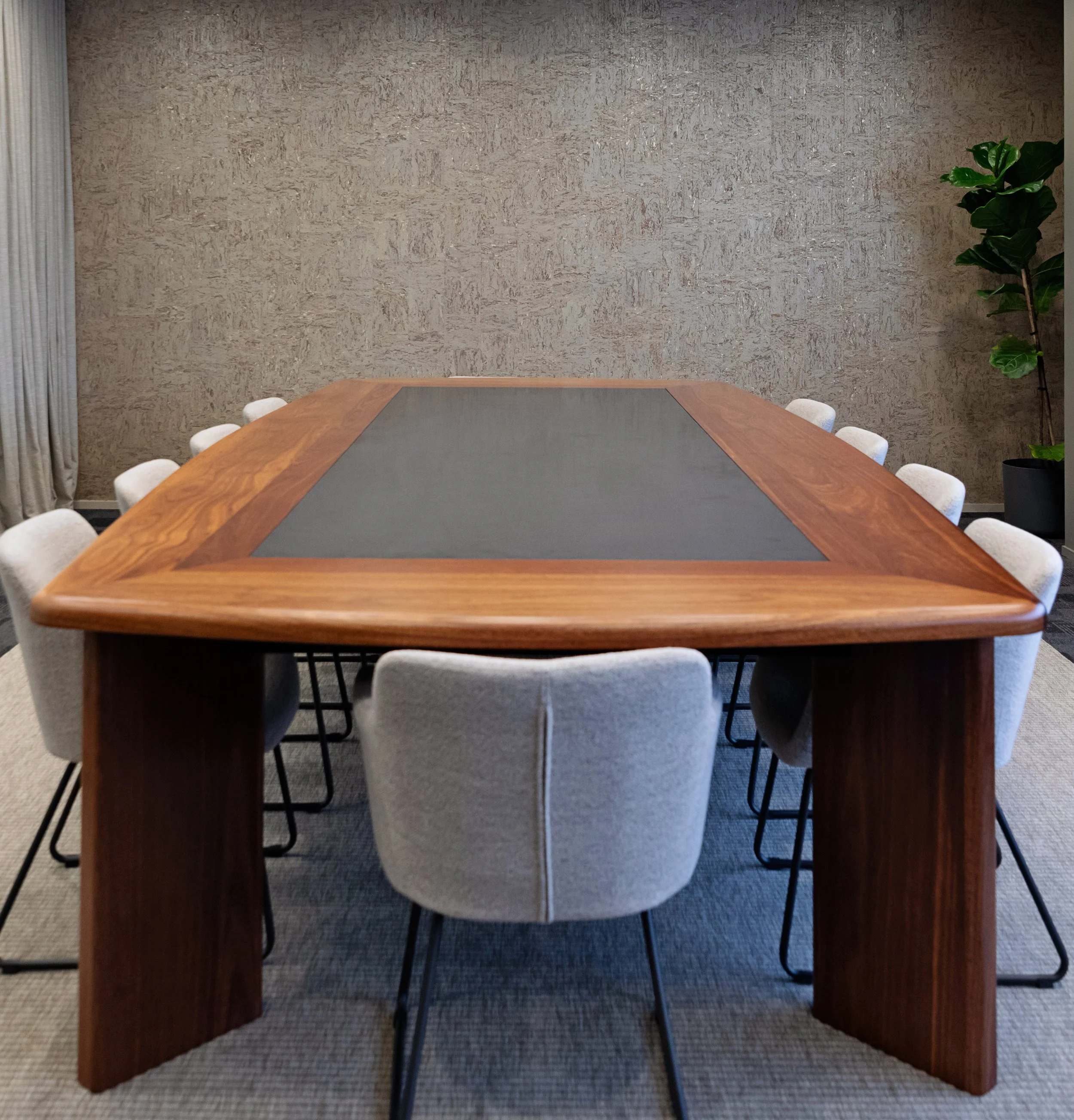 A large wooden conference table with a dark gray center panel, surrounded by white cushioned chairs, in a room with textured beige walls, a gray carpet, a window with vertical blinds, and a tall green potted plant in the corner.