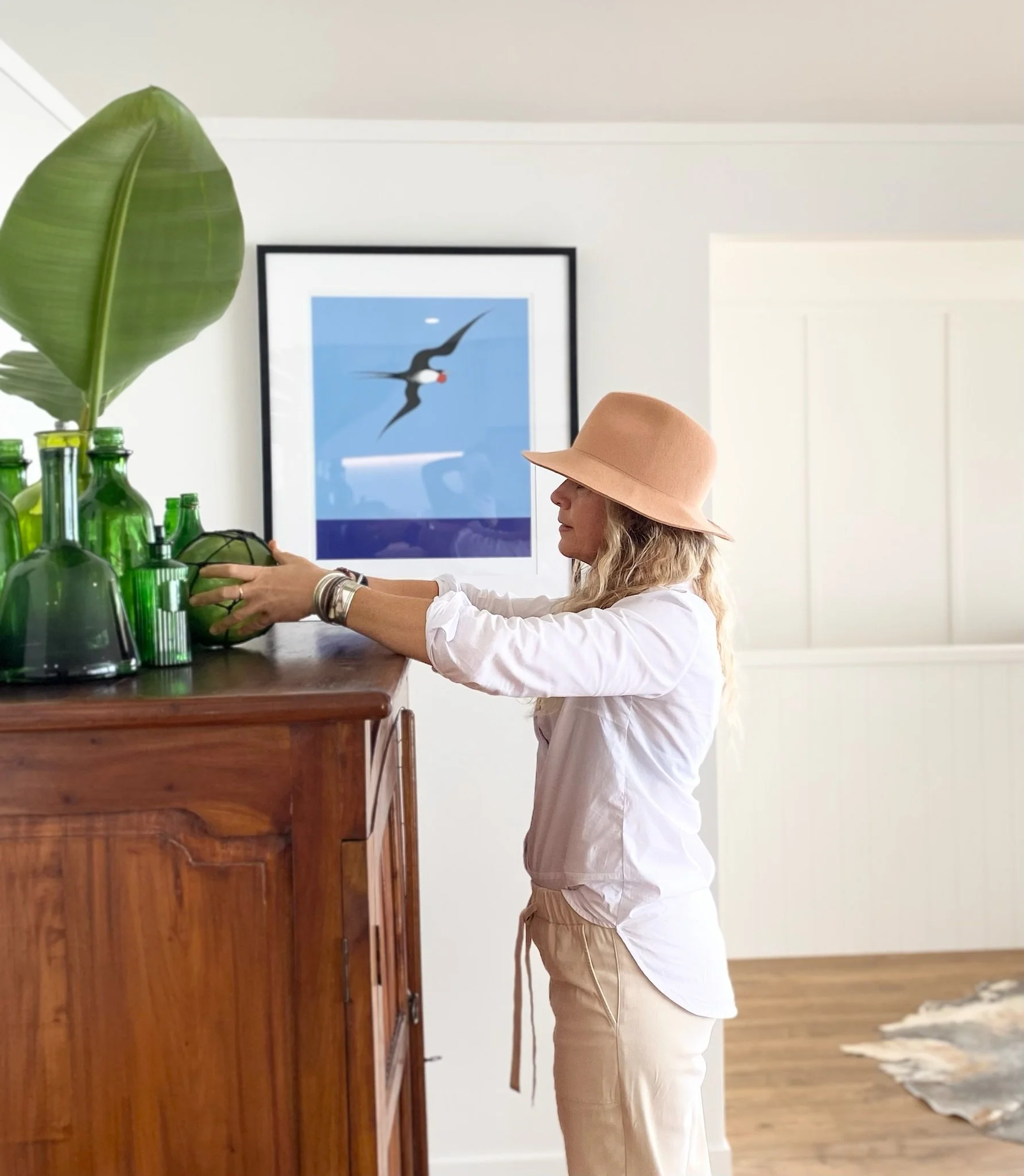 A woman wearing a peach-colored hat and beige pants stands at a wooden counter, reaching for green glass bottles. Behind her is a framed picture of a bird in flight against a blue sky.