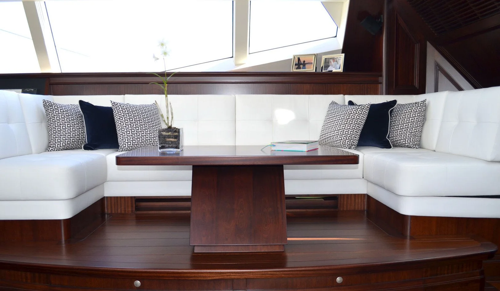 A built-in white leather sofa with a wooden base and a dark wooden coffee table in the center. The sofa has decorative pillows, a plant in a pot, and a few books placed on it. There are picture frames on a shelf behind the sofa, with large windows above allowing natural light into the room.