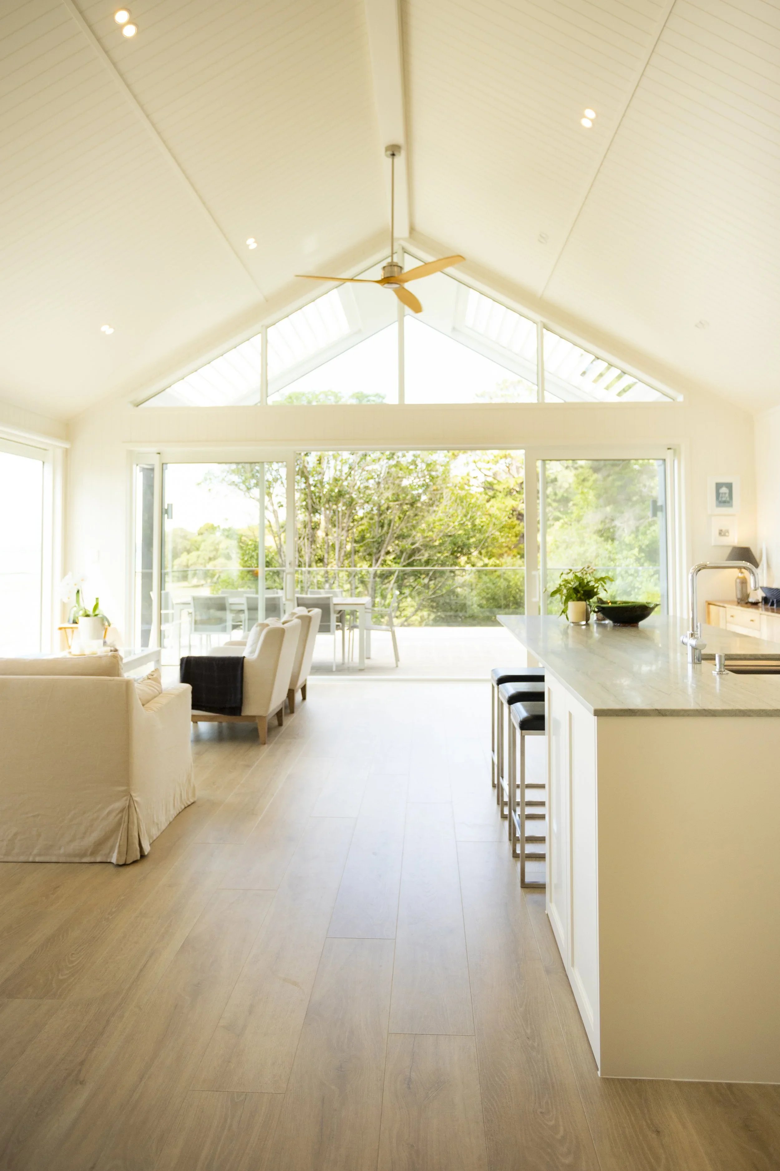 Bright living area with large windows, ceiling fan, and light-colored wooden flooring, connected to a kitchen island with bar stools.