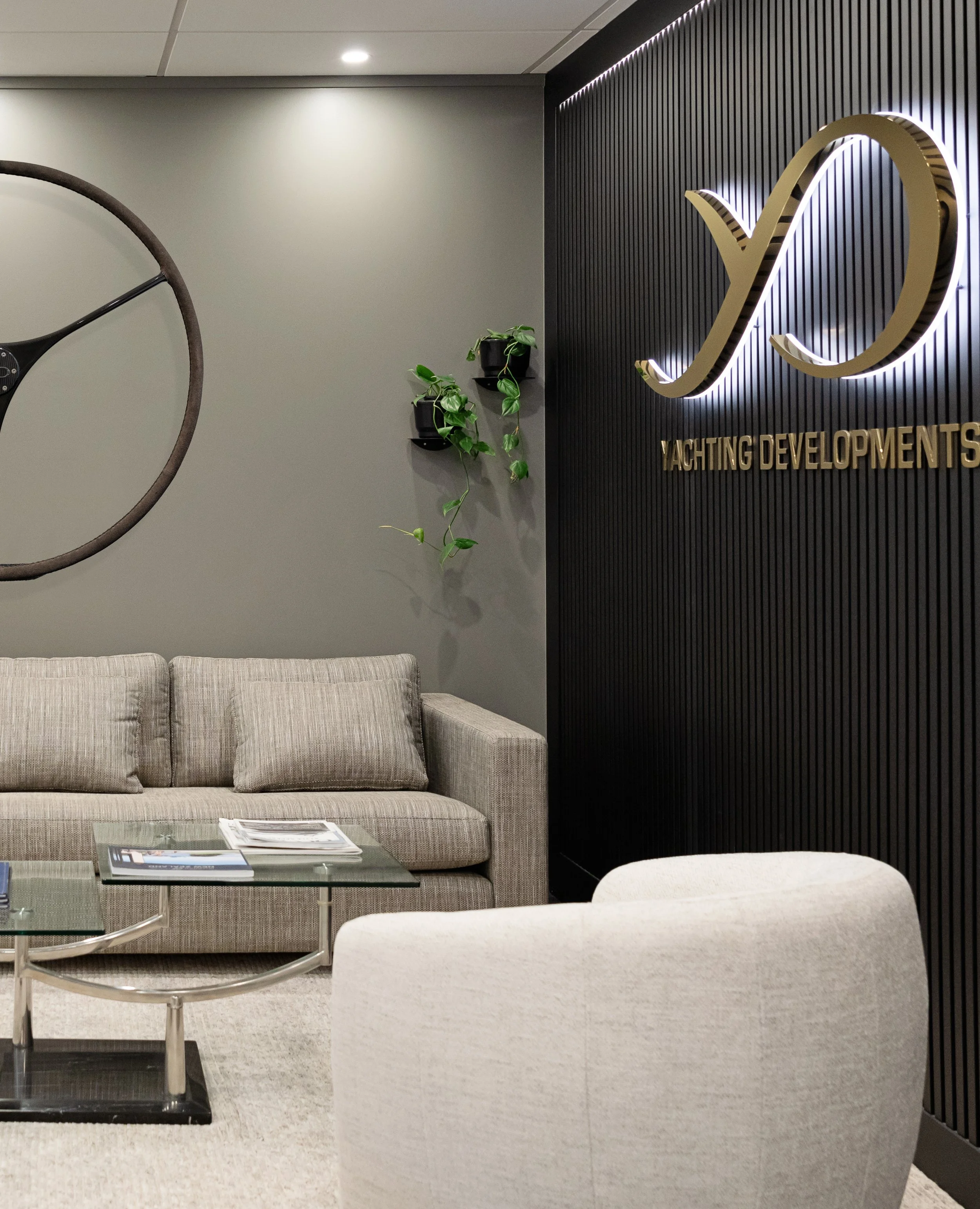 Modern office lounge area with grey couch, glass coffee table with magazines, and black and white decor including a large logo on the wall reading 'Yachting Developments'.
