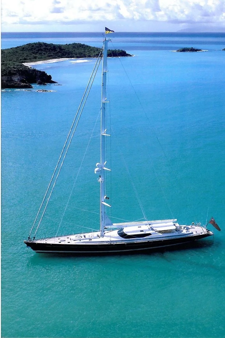 A large yacht with a tall mast anchored in clear blue water near a lush green island with a sandy beach.