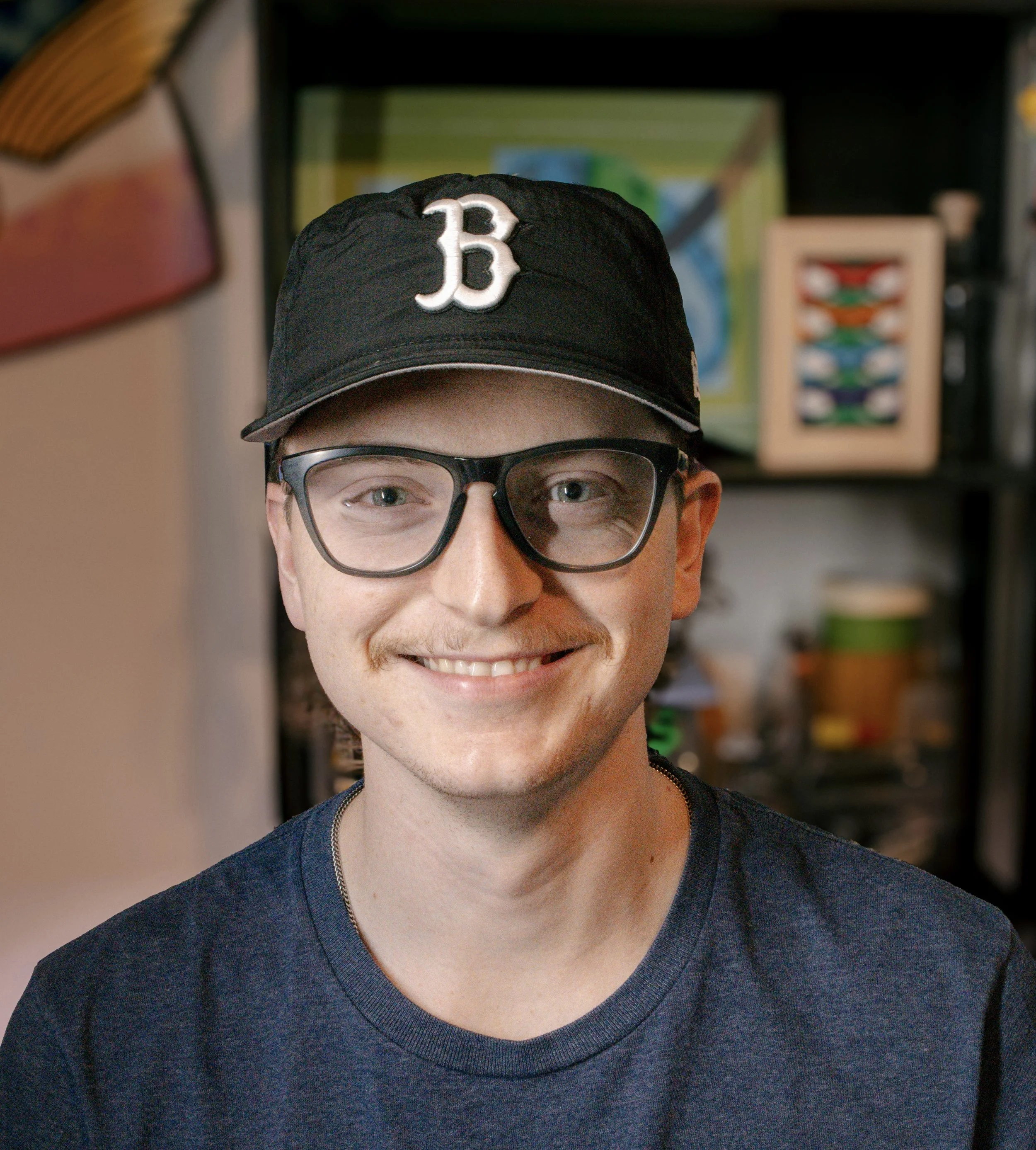 A young man with glasses and a mustache smiling, wearing a black baseball cap with a white 'B' on it, in front of a bookshelf.