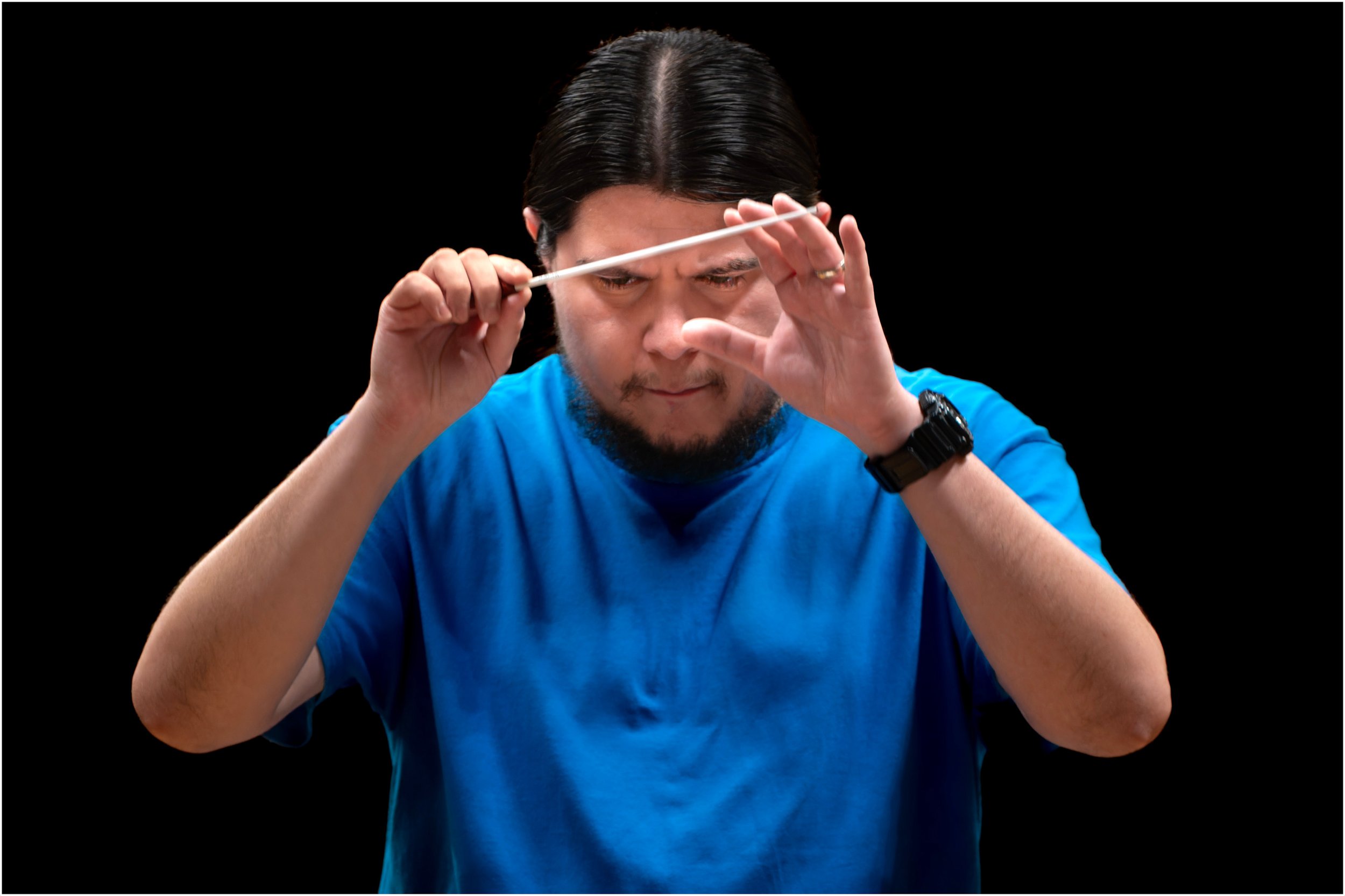 Man with long dark hair, wearing a blue shirt and black watch, conducting an orchestra with a baton against a black background.