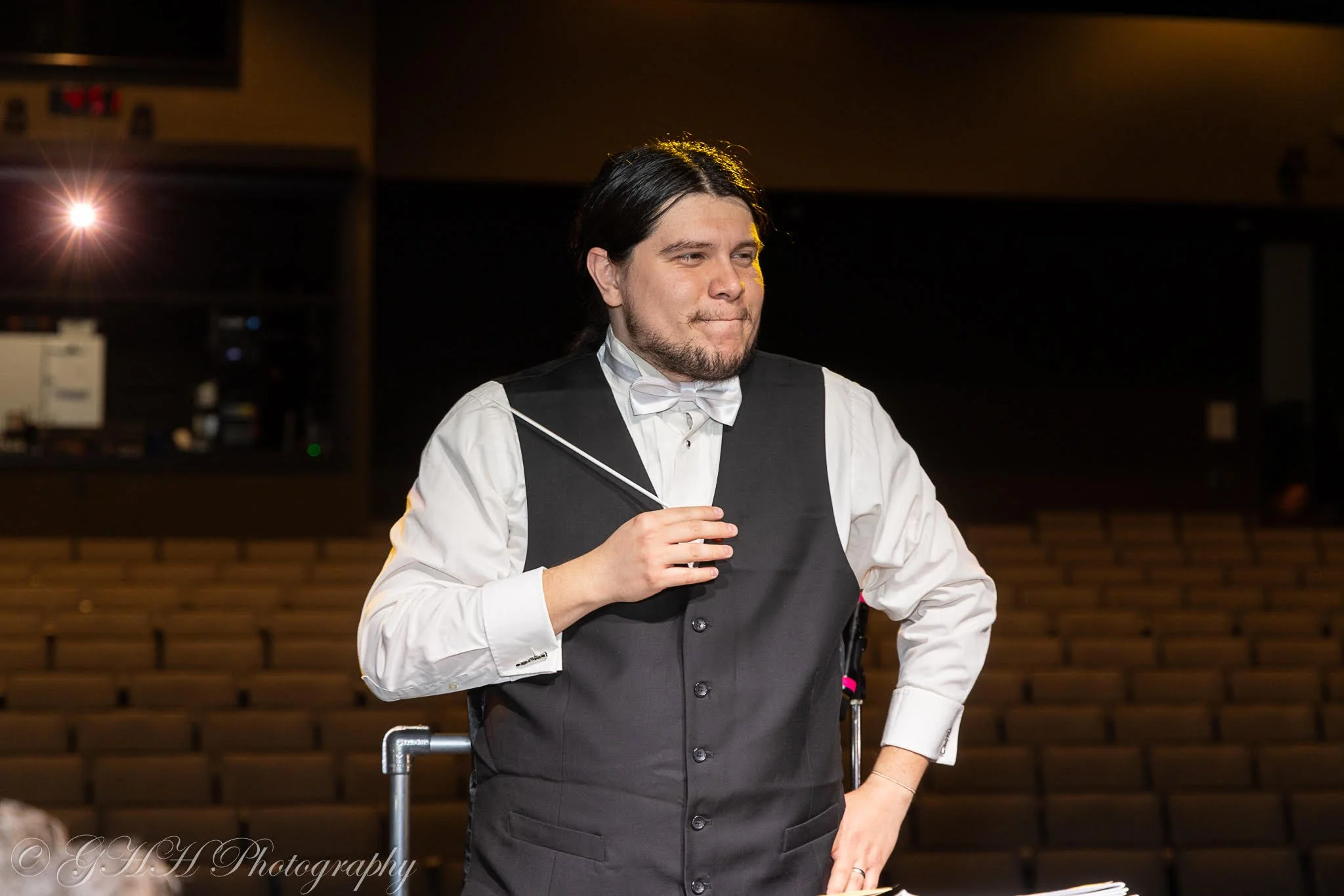 A conductor wearing a white dress shirt, black vest, and bow tie holding a baton in his right hand during a performance in a theater auditorium.