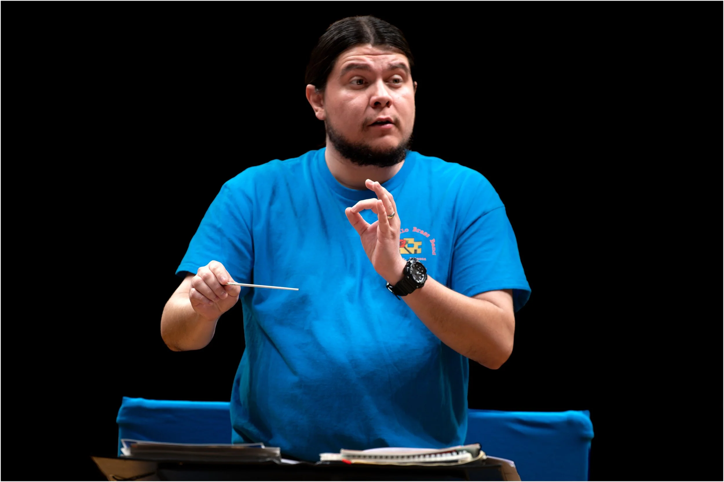A man in a blue T-shirt is conducting a music rehearsal or class, holding a baton in his right hand and gesturing with his left hand, set against a black background.