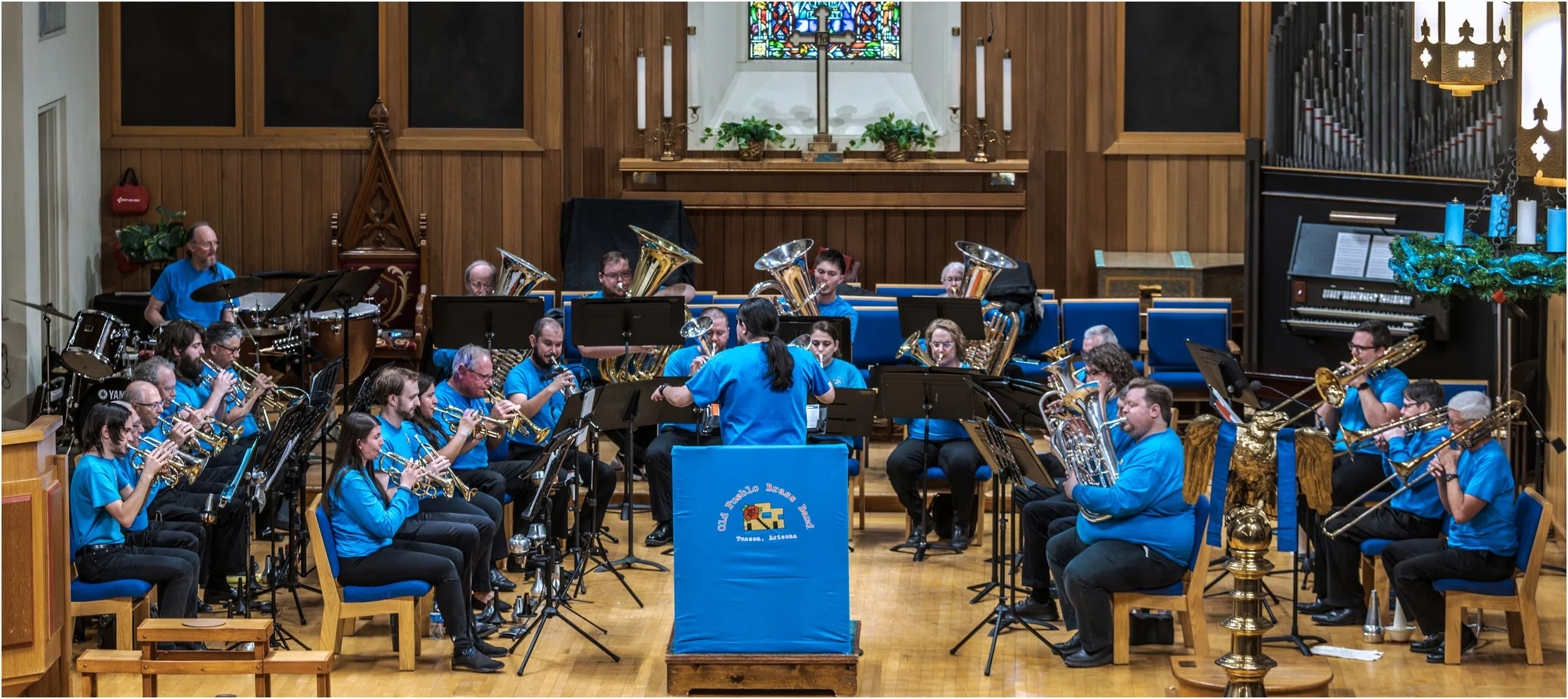 A large brass band performing in a church with wood-paneled walls and stained glass windows. The band members are wearing matching blue shirts and playing various brass and percussion instruments. There are musical stands and a conductor leading the group.