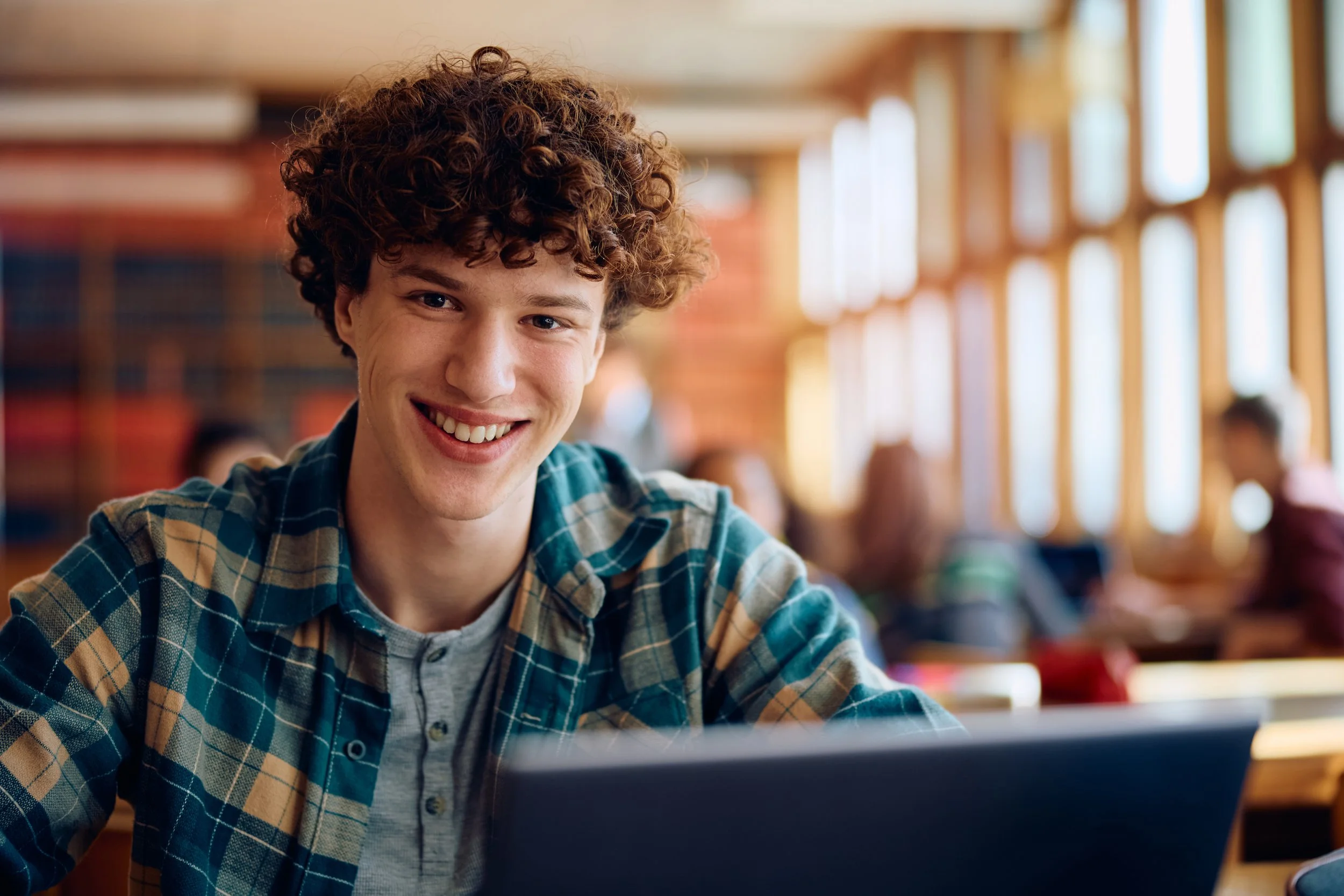 A young man with curly hair and a checkered shirt smiling at the camera while working on a laptop in a bright, modern classroom or library.