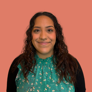 A woman with curly hair wearing a teal blouse with a black cardigan, smiling against a pink background.