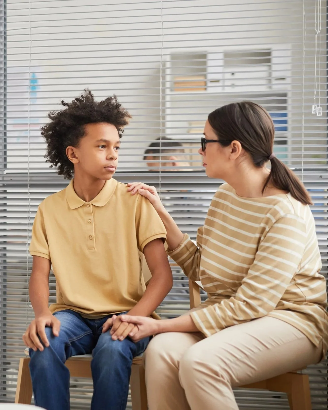 A woman from the Family Urgent Response System program with glasses and a striped beige shirt speaks with a young boy with curly hair, wearing a yellow polo shirt, in a room with glass blinds.