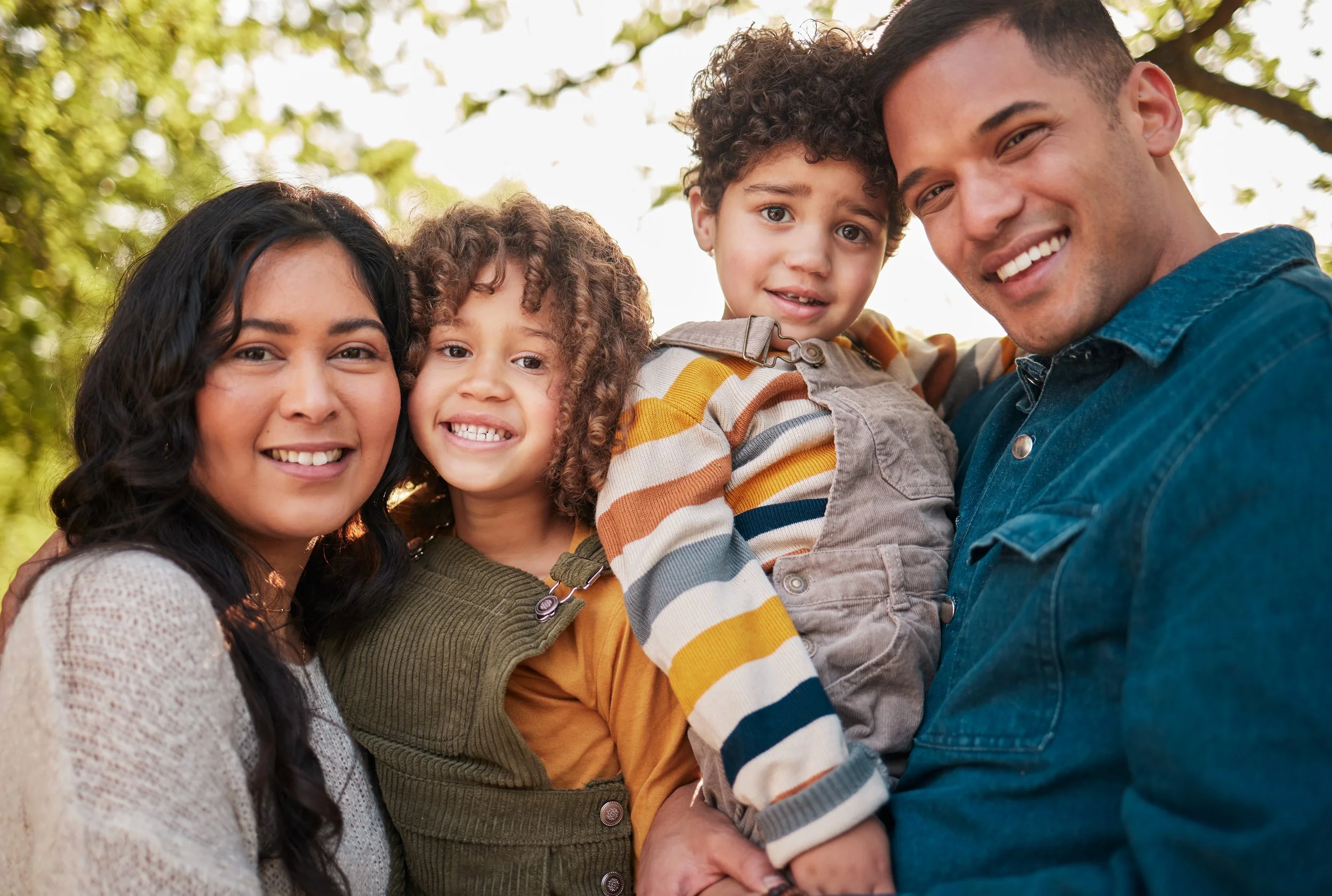 A happy family of four, including two foster kids, smiling together outdoors, with trees and sunlight in the background.