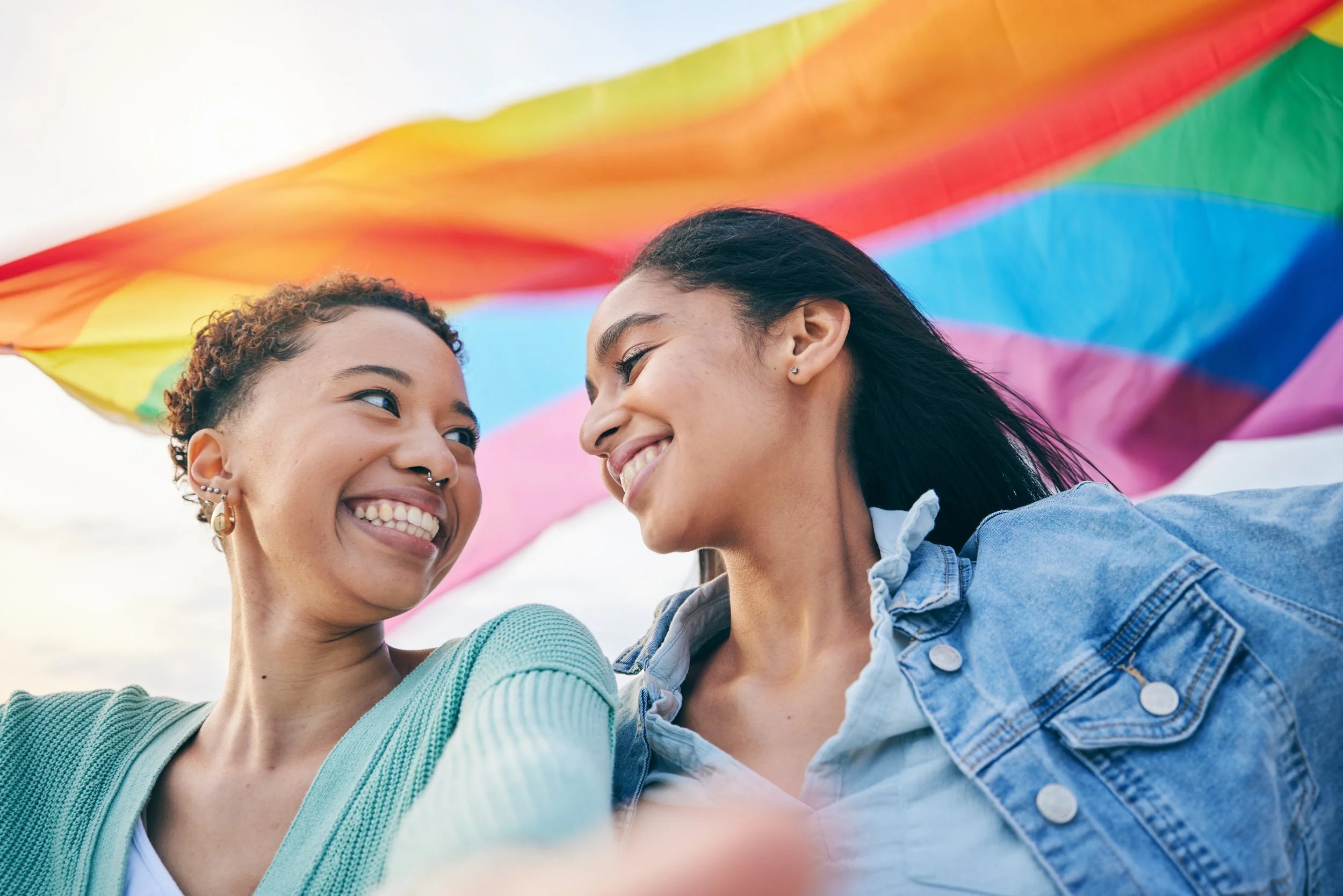 Two smiling women looking at each other under a rainbow pride flag.