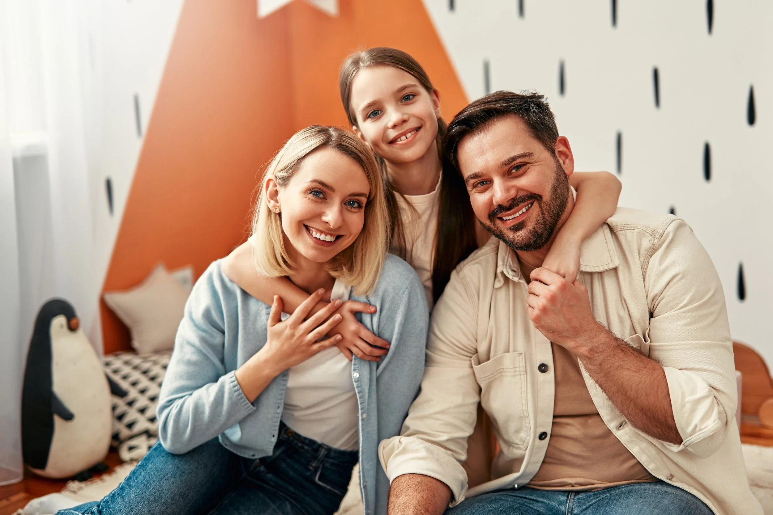 A foster family of three smiling people, a woman, a man, and a girl, sitting together in a cozy room with playful decor, including penguin plush toy and geometric patterned pillow, during daytime.