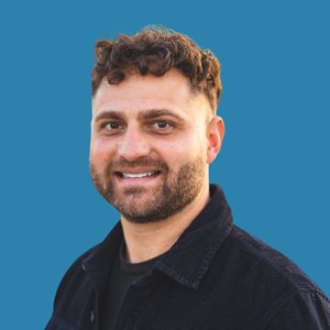 Portrait of a smiling man with curly brown hair and a beard, wearing a dark jacket against a blue background.