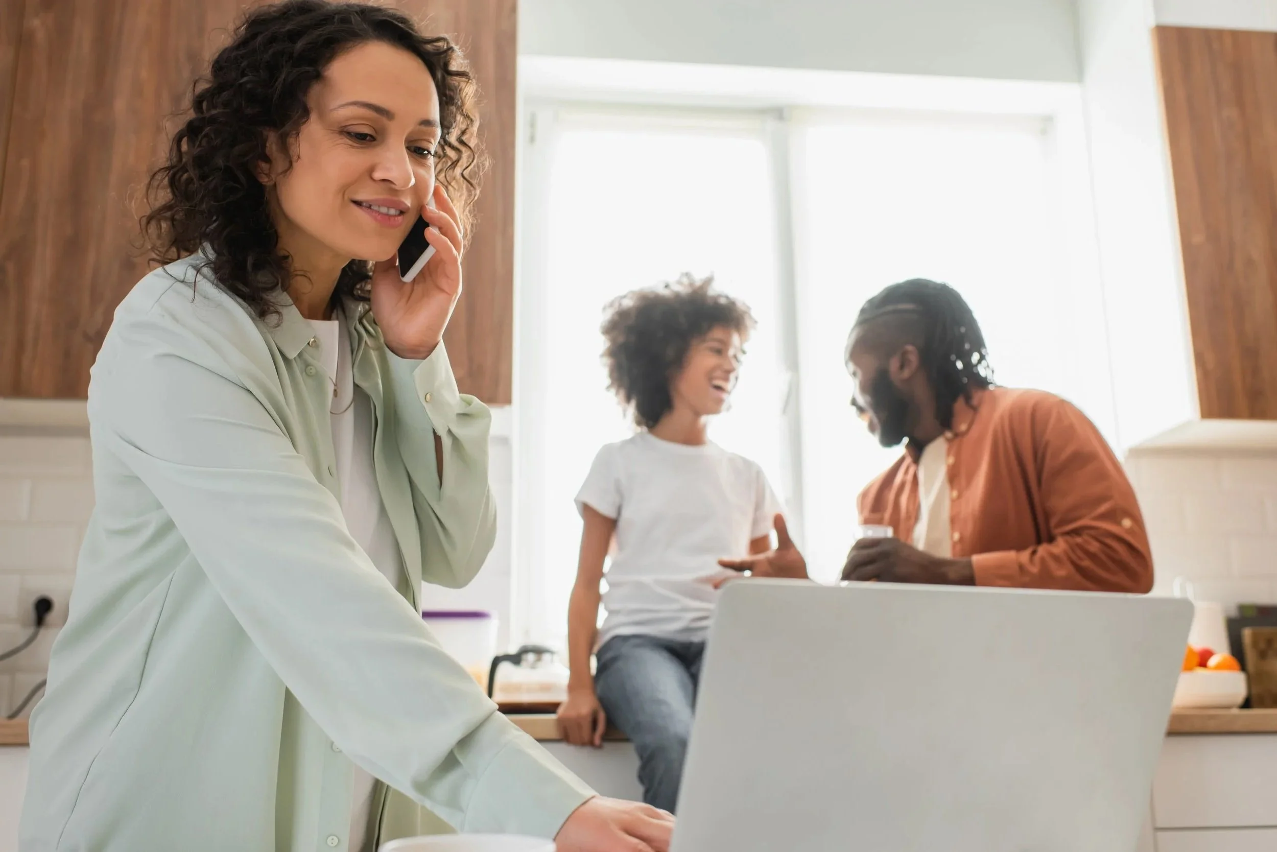 Three family members in a modern kitchen. One talking on the phone and on a laptop reaching out to the Family Urgent Response System.