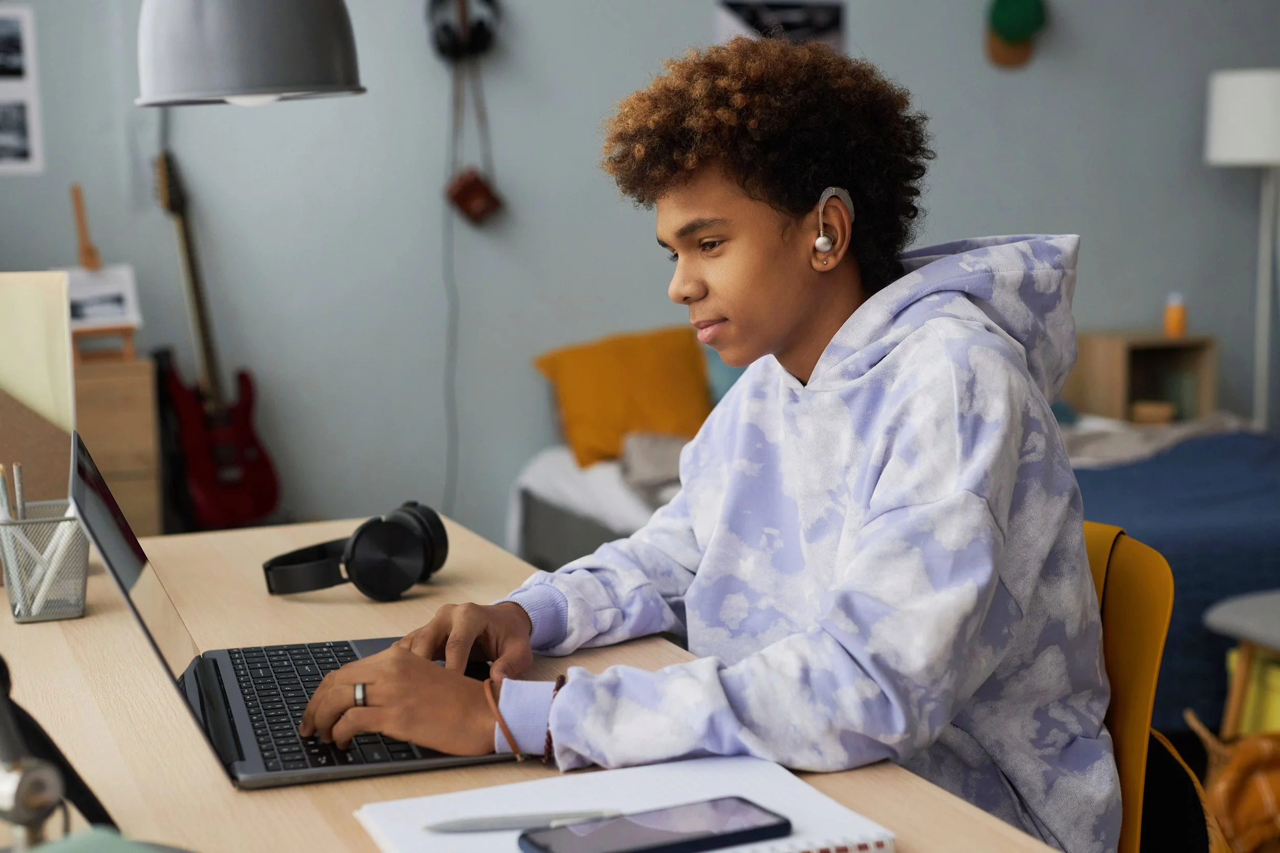 A young person sitting at a desk, working on a laptop, wearing headphones, in a bedroom with a bed, guitar, and various items in the background.