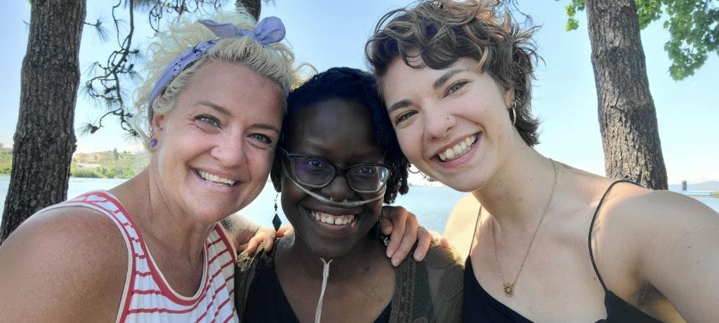 Three women standing outdoors near a body of water, smiling for a selfie.