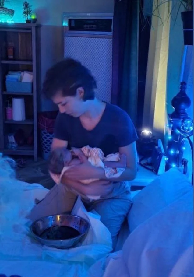 A person with short dark hair holding a newborn baby wrapped in a blanket, sitting on a bed in a dimly lit room with blue lighting.