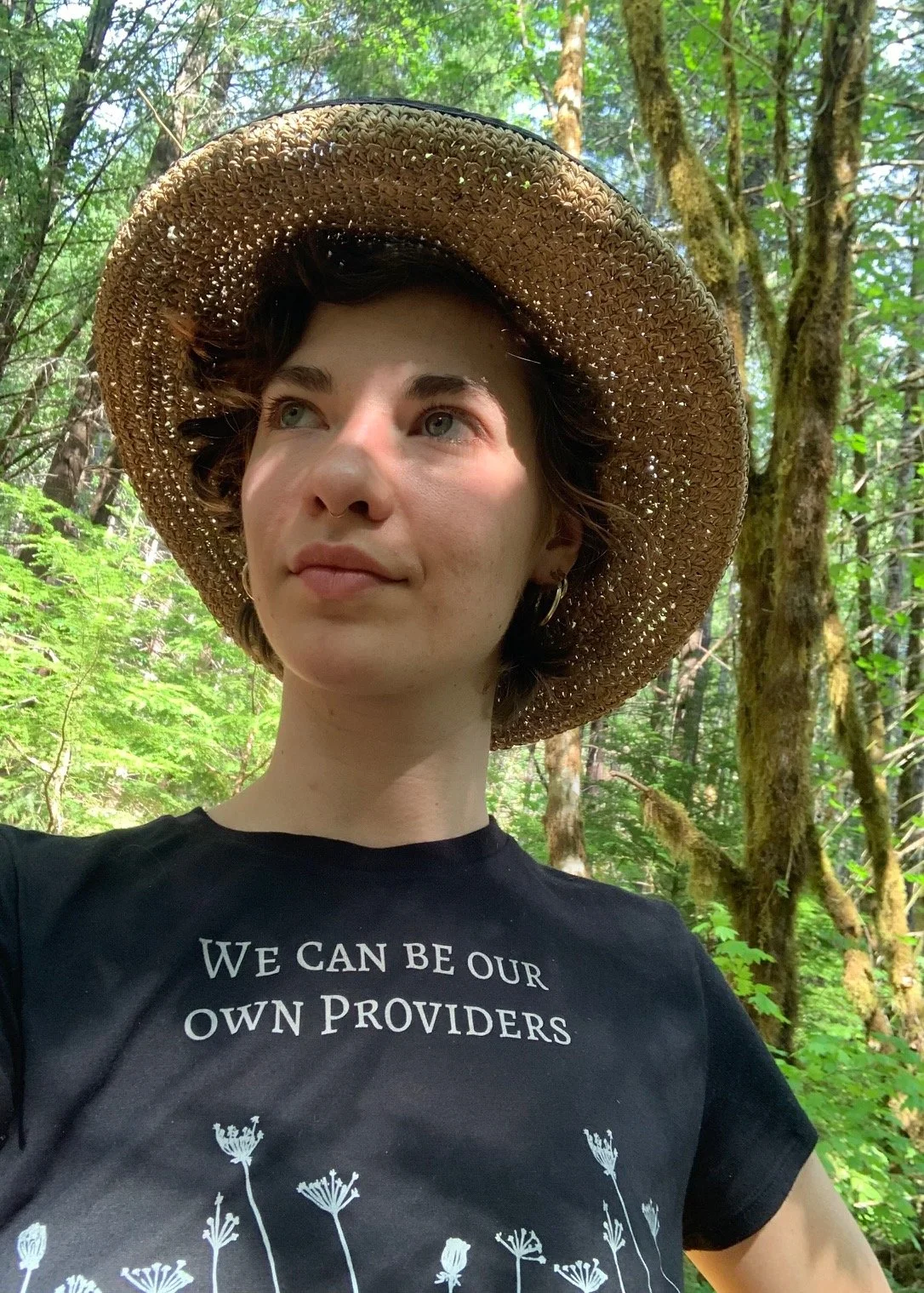 A young woman with short brown hair, wearing a large straw hat and black t-shirt with a floral design and the phrase 'We can be our own providers,' stands in a lush, green forest.