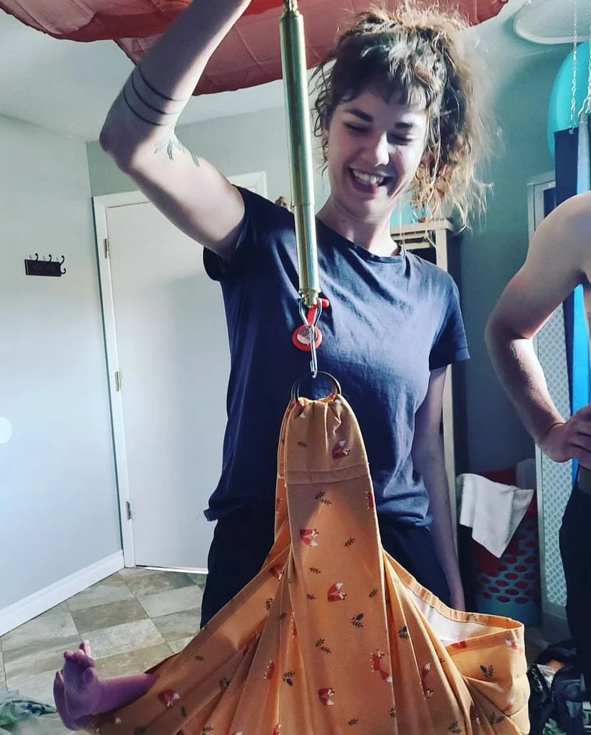 A woman with curly hair, smiling, is holding a scale with a fabric bag hanging from it. The fabric bag has a deer print and appears to contain a large, pink plush toy. The woman is in a room with light-colored walls and various household items in the background.