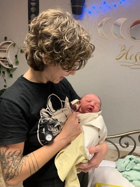 Young woman with curly hair holding a newborn baby wrapped in a blanket in a hospital room.