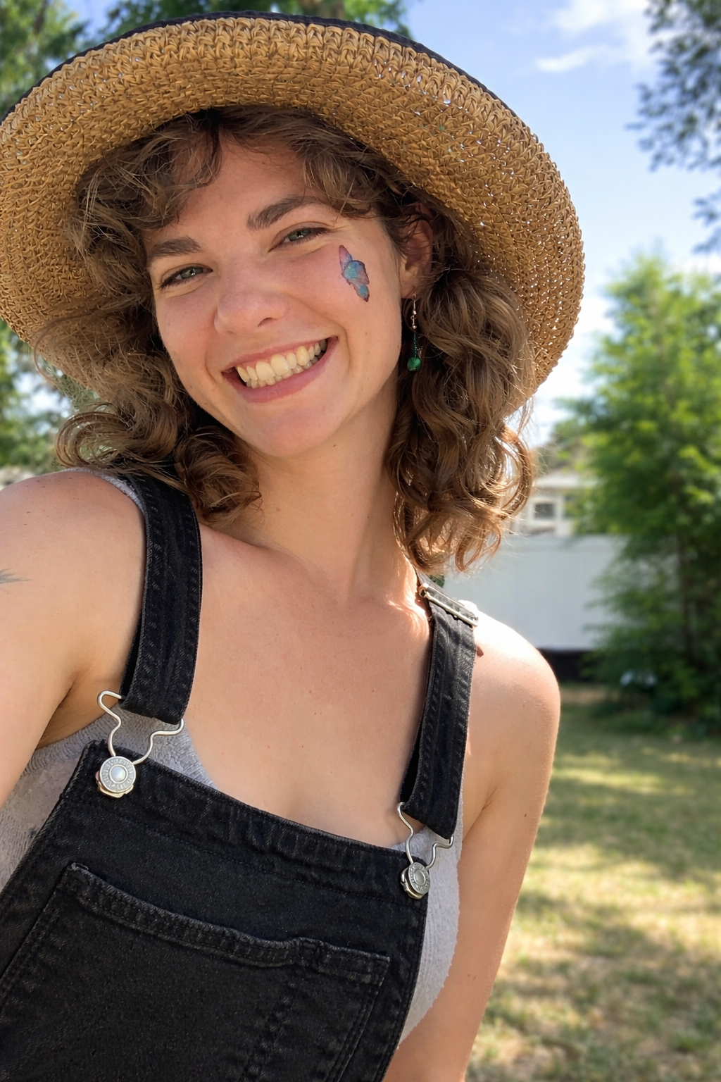 A young woman smiling outdoors wearing a wide-brimmed straw hat, black overalls, and a white tank top. She has a blue and purple butterfly tattoo on her cheek, green dangle earrings, and curly light brown hair. The background has green trees and a small white building.