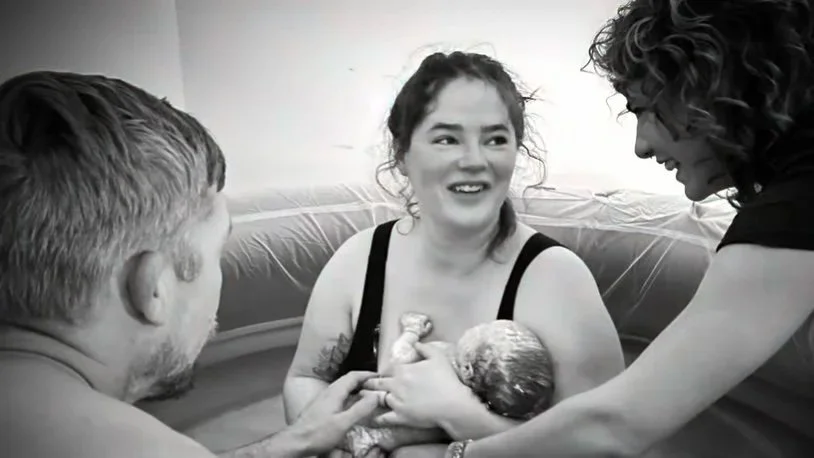 Woman holding newborn baby during water birth, surrounded by her partner and midwife in a birthing pool.