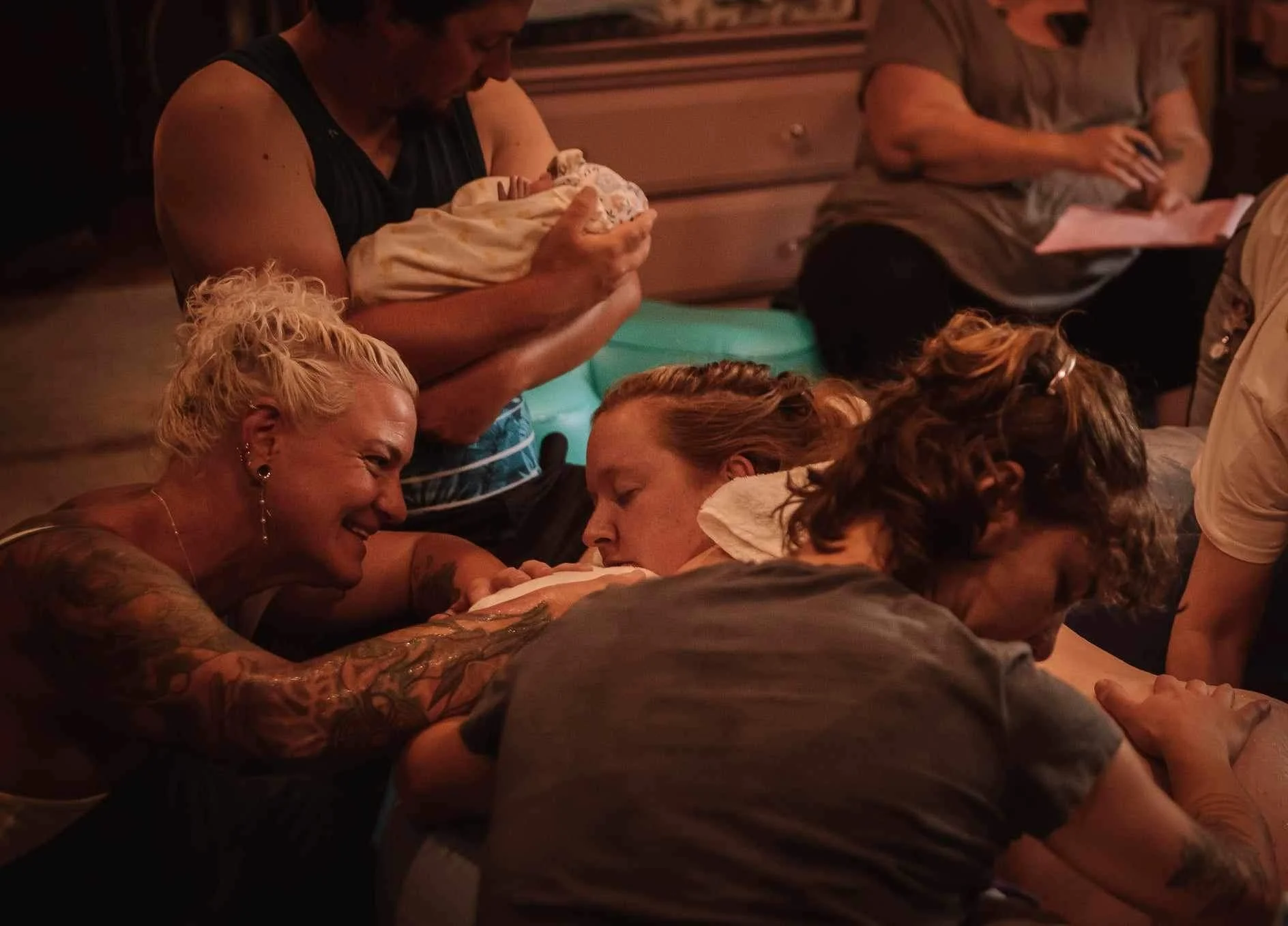 Group of people, including a woman with tattoos and short blonde hair and a woman with reddish hair, engaging in a close, possibly intimate, activity on a table in a dimly lit room. One woman is holding a baby.