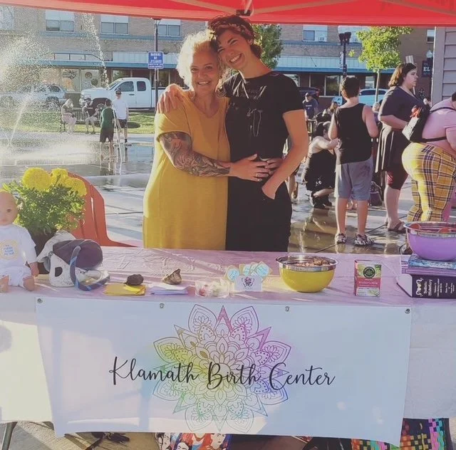 Two women smiling and hugging at a booth with a banner that reads "Klamath Birth Center"; outdoor event with people and a fountain in the background.