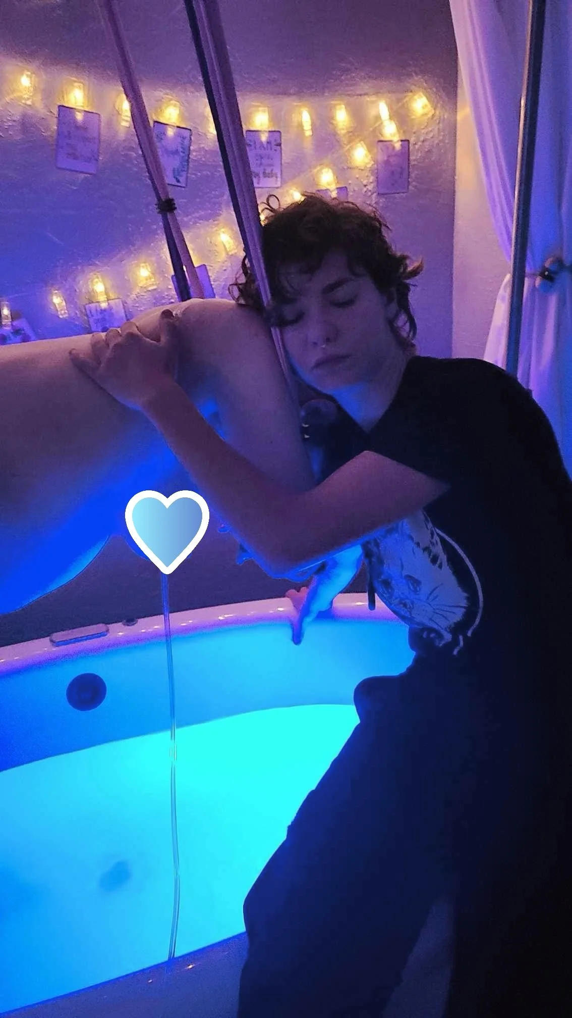 A person with curly hair and a dark shirt hugging a dolphin on a blue-lit boat, with string lights and framed pictures in the background.