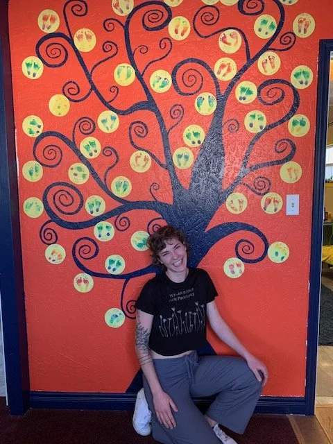 A woman with curly hair and tattoos crouches in front of a painted wall featuring a tree with black swirling branches and yellow circular fruit or leaves, with some leaves decorated with green and red designs, on an orange background.