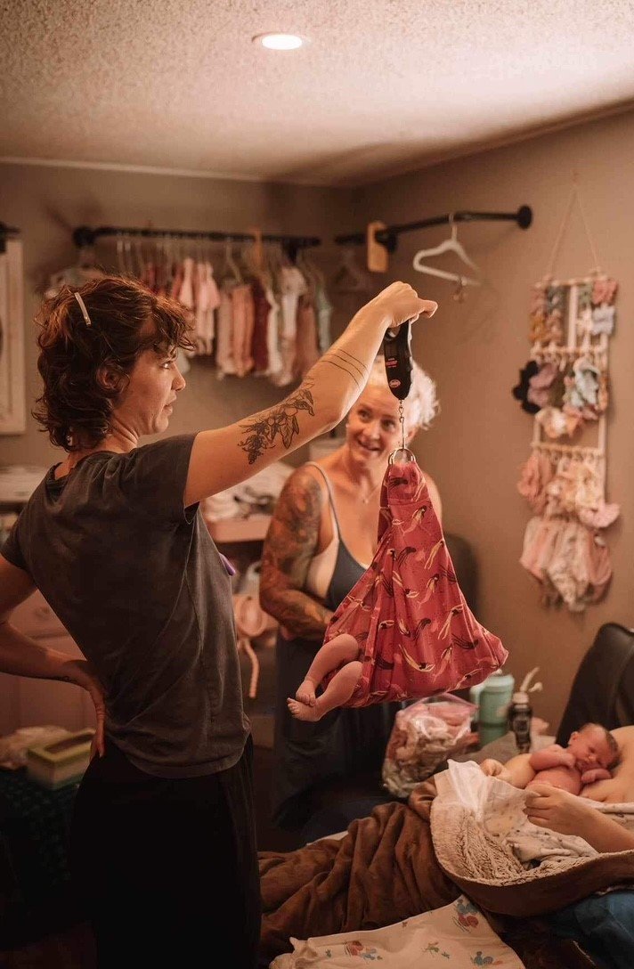 Two women and a baby in a bedroom, with clothing and sock displays on the wall. One woman holds a pink baby dress with hanging baby socks.
