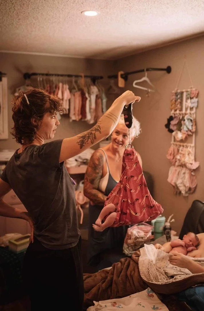 Two women in a nursery or bedroom with a baby lying in a crib. One woman is holding a small pink dress, and the other woman is smiling, looking at the dress.