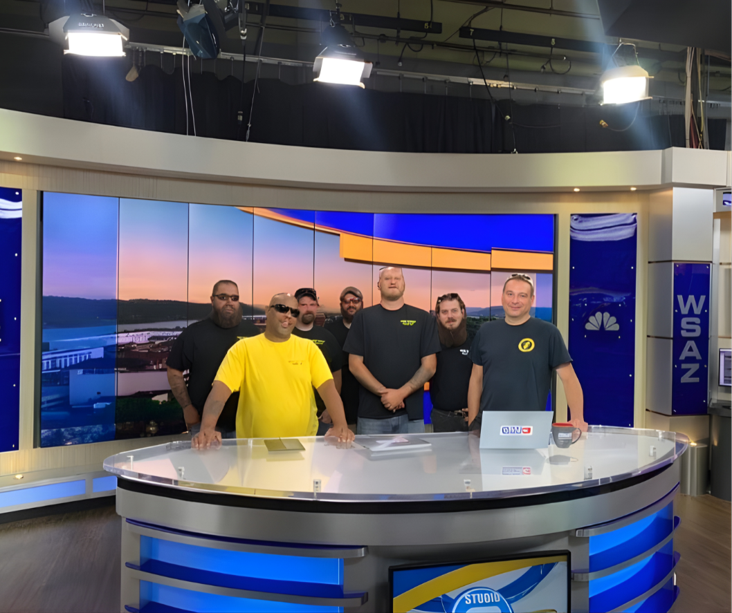 Six men standing behind a news studio desk with a large screen displaying a sunset over a river in the background.