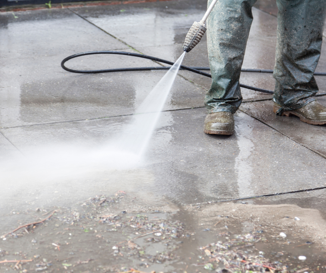 Person in work boots pressure washing a concrete sidewalk on a rainy day.