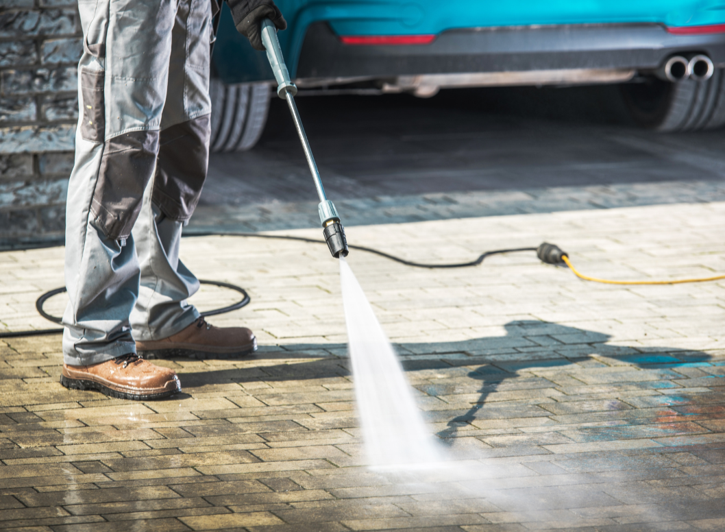 Person cleaning brick pavement with a pressure washer near a blue car.
